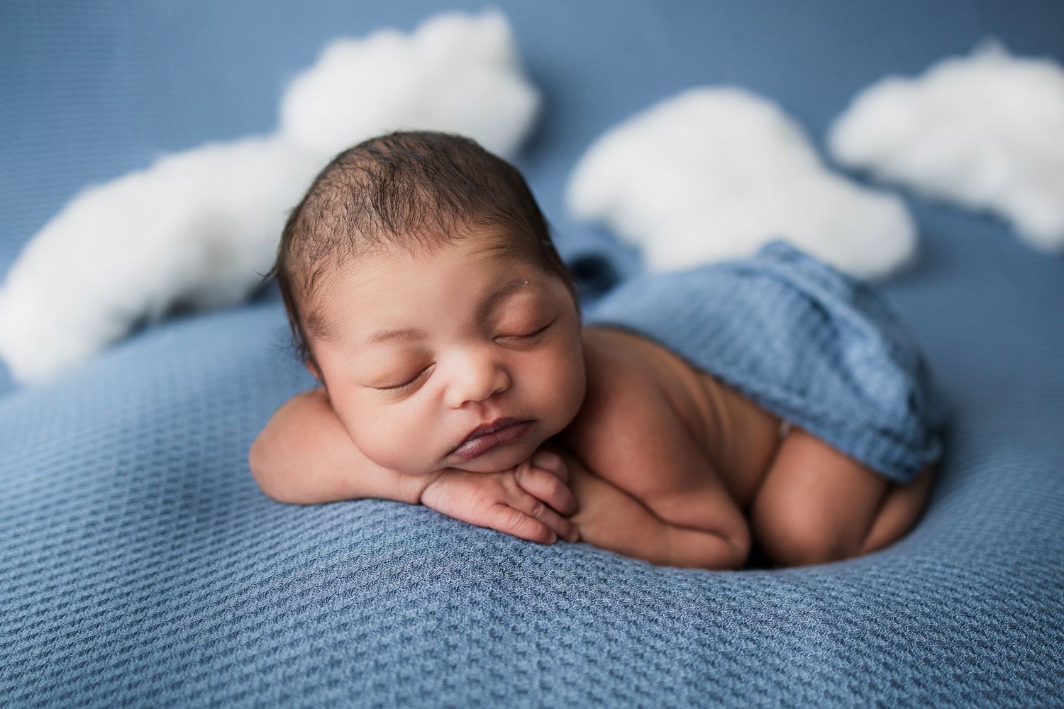 A sleeping newborn nestles on a blue blanket with decorative cotton clouds arranged above.