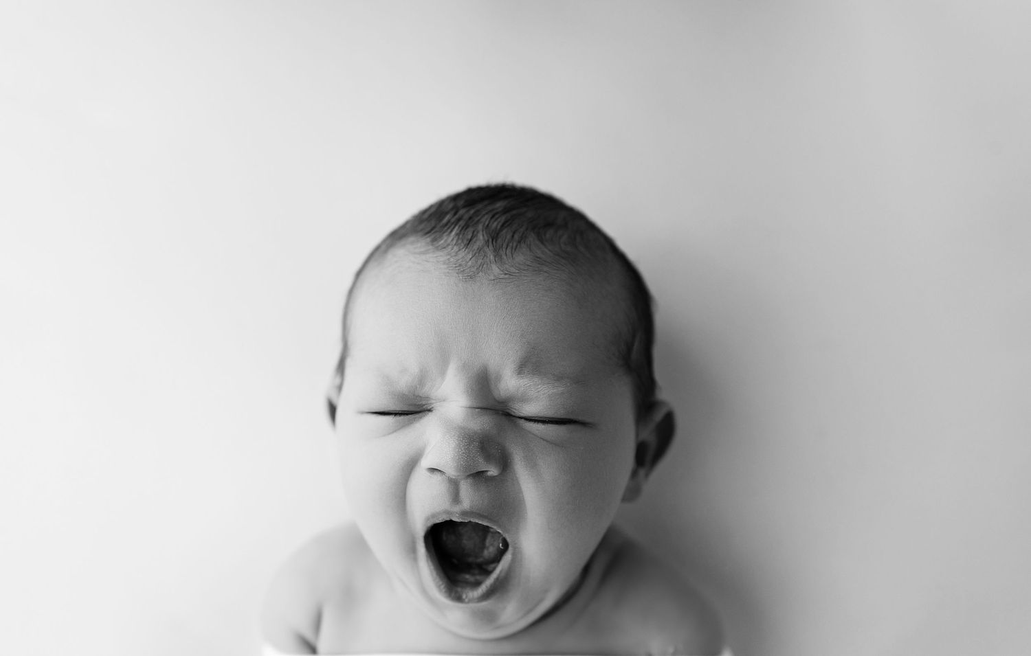 Black and white photo of a newborn baby yawning with eyes closed against a white background.