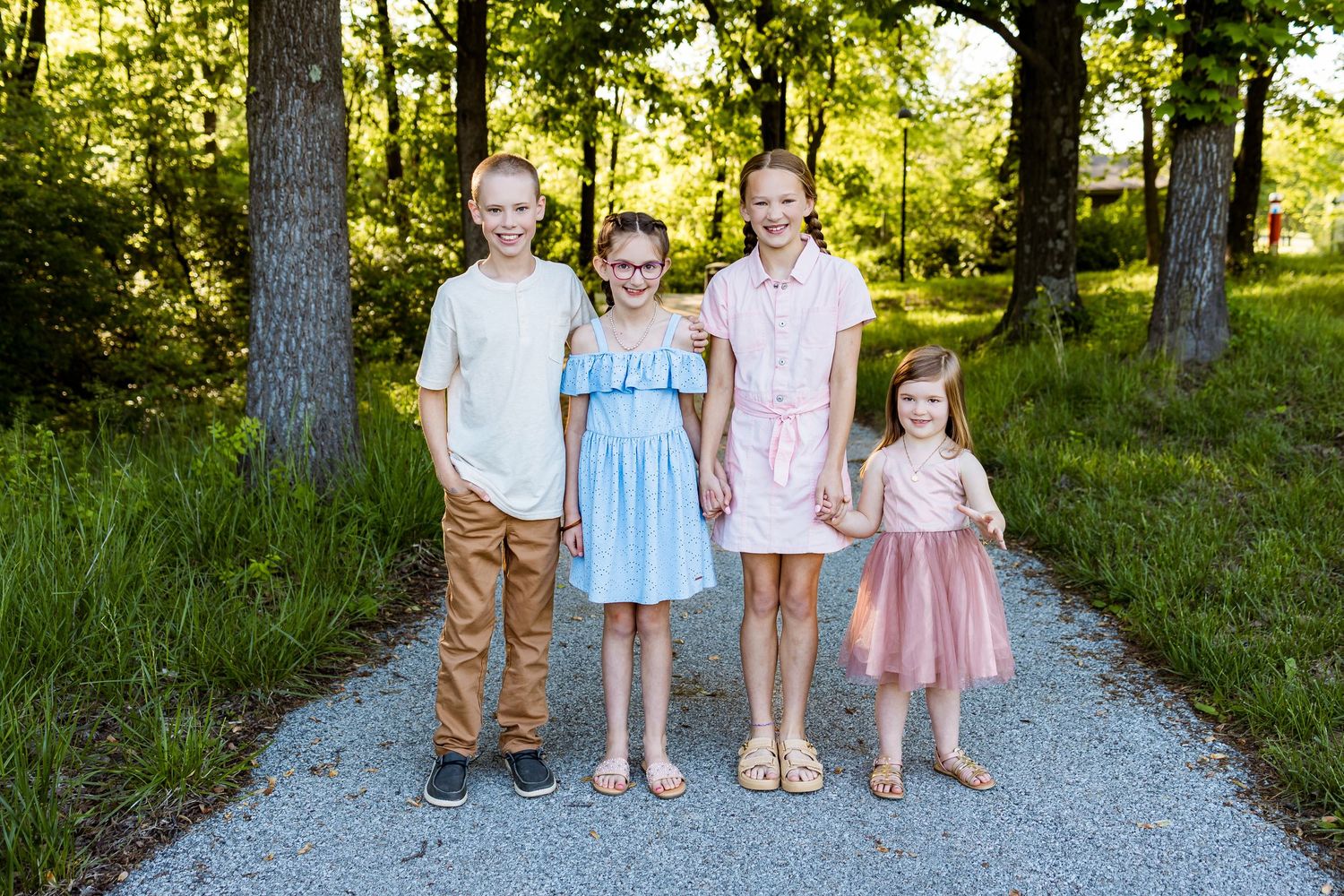 Four children standing together on gravel path with sunlit forest backdrop.