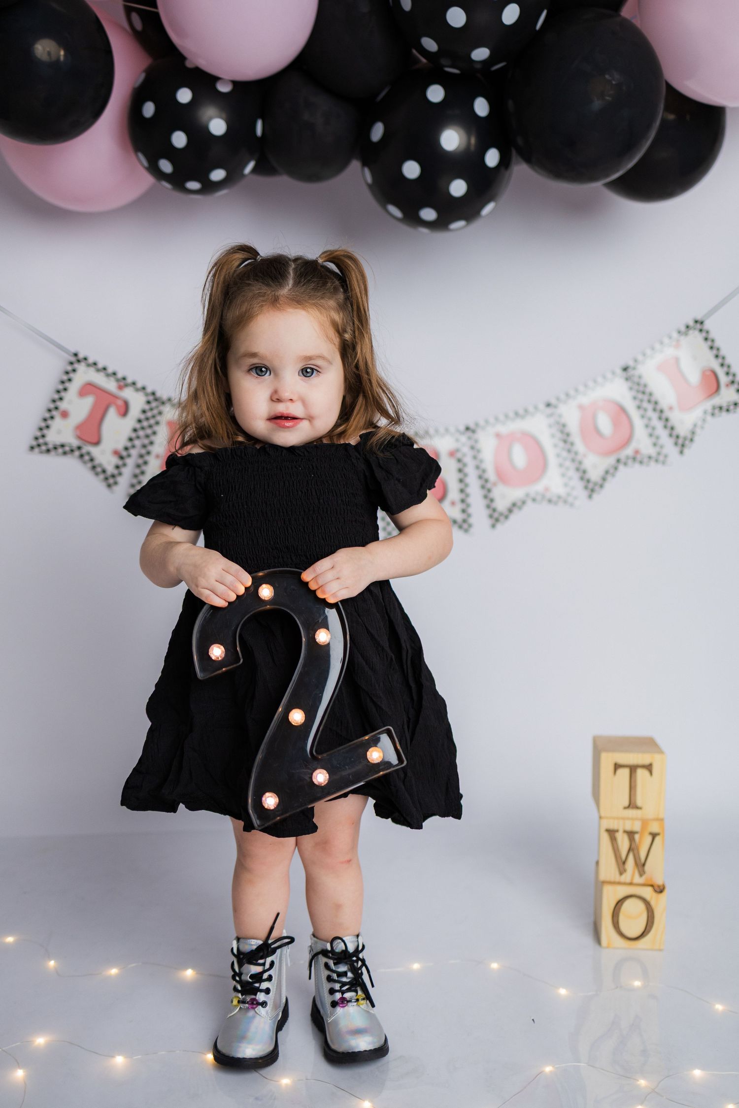 Birthday celebration setup with black and pink polka dot balloons, silver letter banner, and wooden number blocks showing 'TWO'.