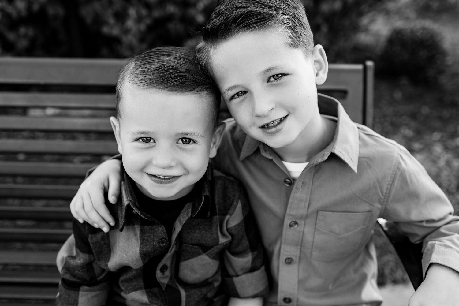 Two children smile and embrace while sitting together on a wooden bench outdoors.
