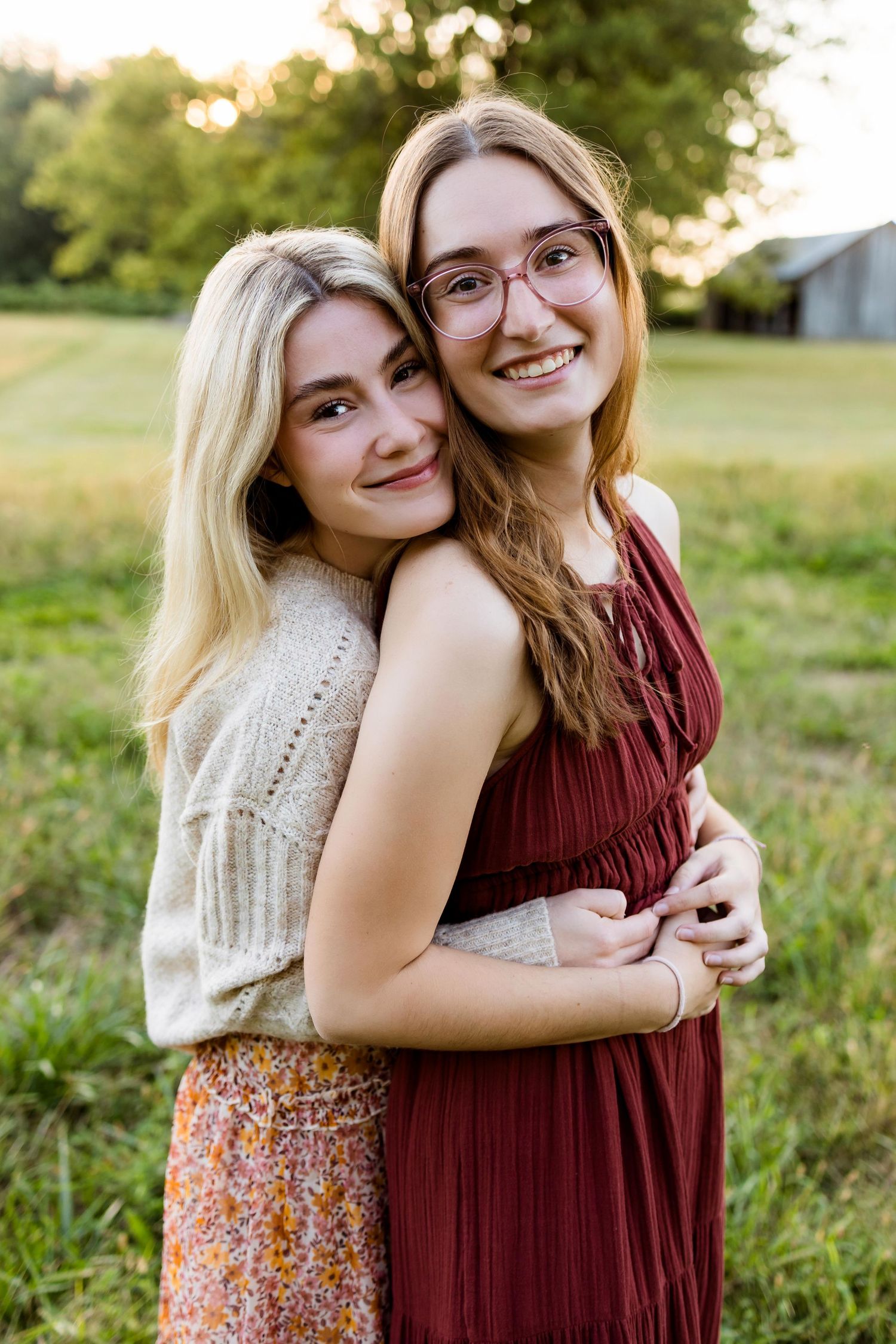 Two friends embrace and smile together in a sunny field wearing burgundy and floral dresses during golden hour.