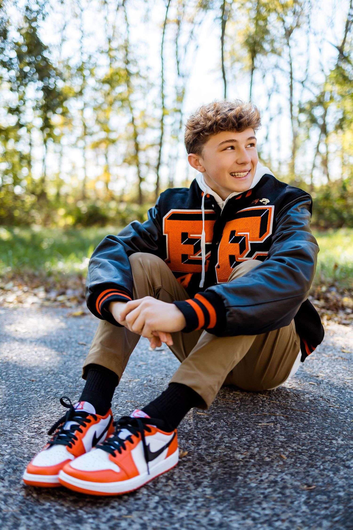 Person wearing varsity jacket and orange sneakers sits casually on gravel path surrounded by autumn trees.