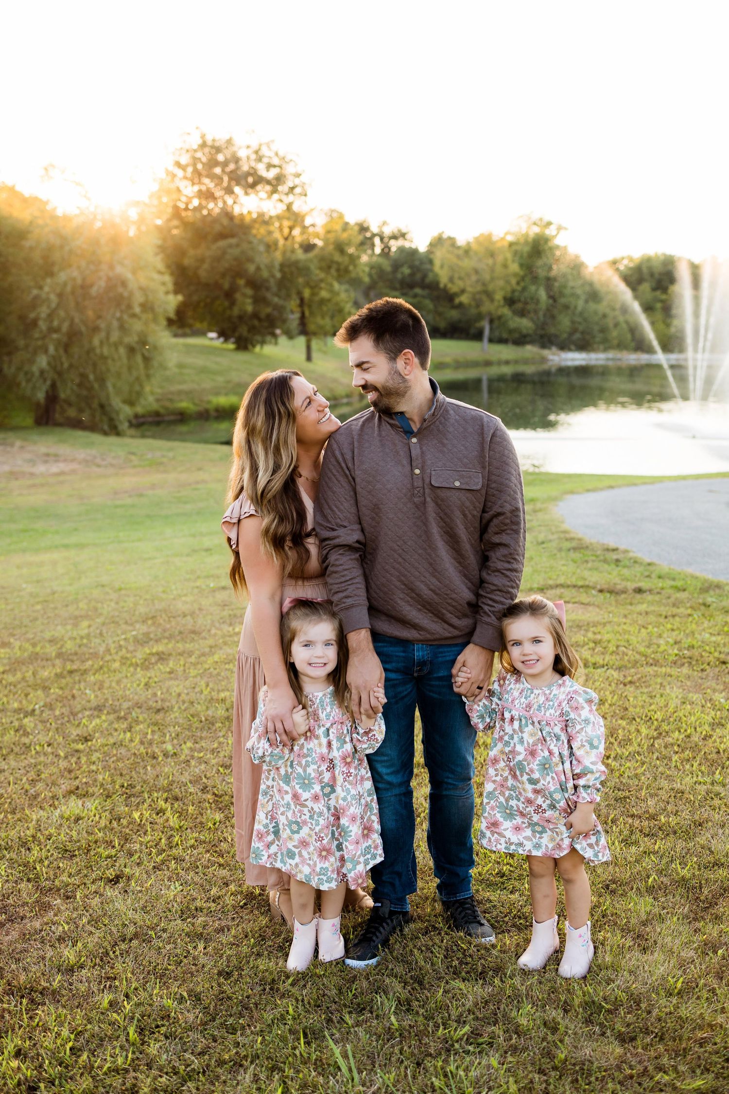 A family stands together on a grassy hill during golden hour with a fountain in the background.