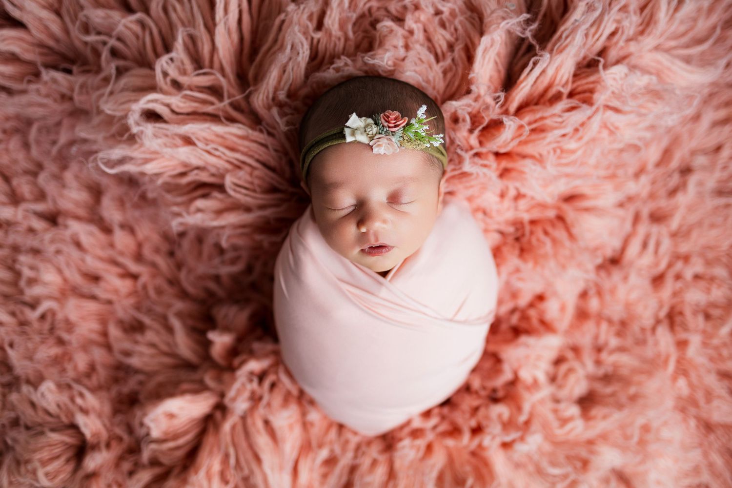A sleeping newborn baby swaddled in white fabric nestled among fluffy peach-colored textured blankets.