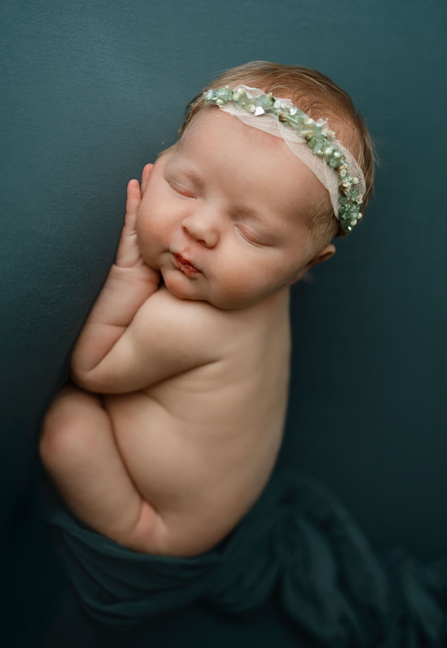 A sleeping newborn wrapped in dark teal fabric wearing a delicate floral headband in a professional studio portrait.