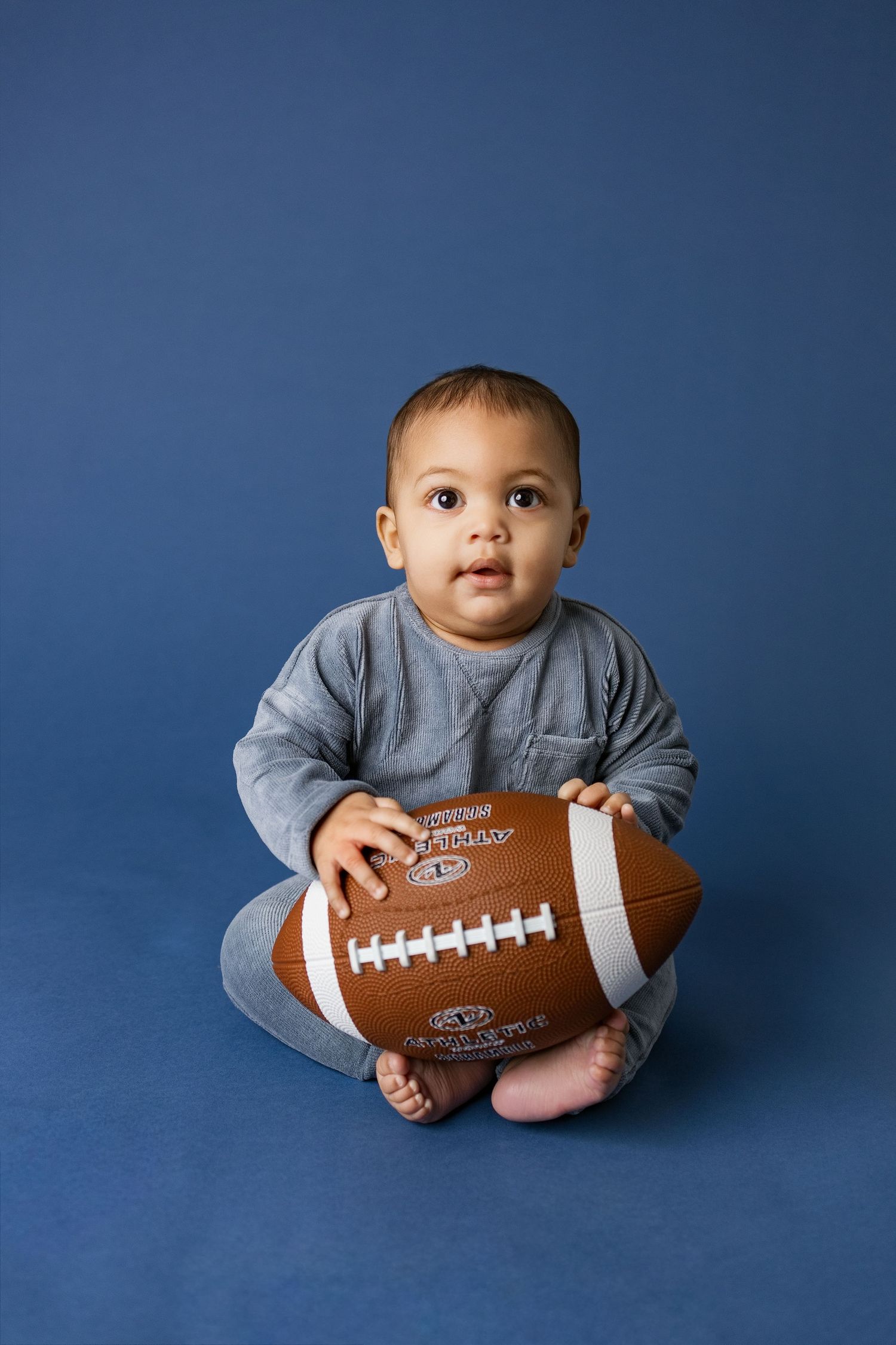 Baby in gray sweater poses with football against blue backdrop in professional studio photo series.