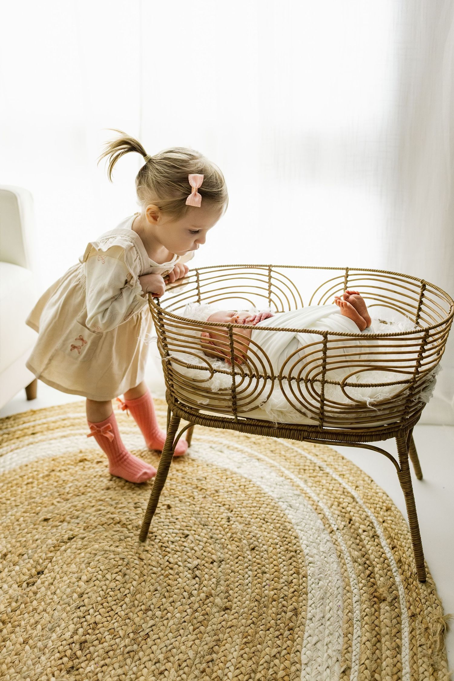 Little girl in white dress exploring vintage rattan furniture on natural jute rug in bright room.