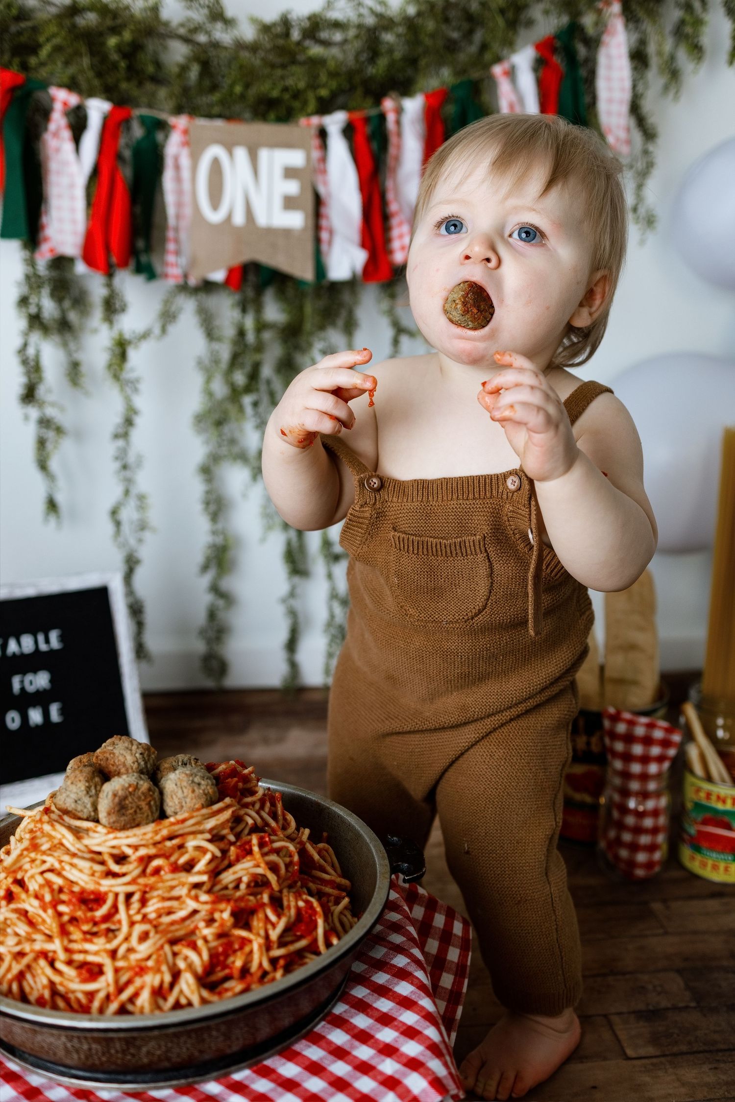 Small child in brown overalls sits at table with spaghetti and meatballs during Italian-themed first birthday party.