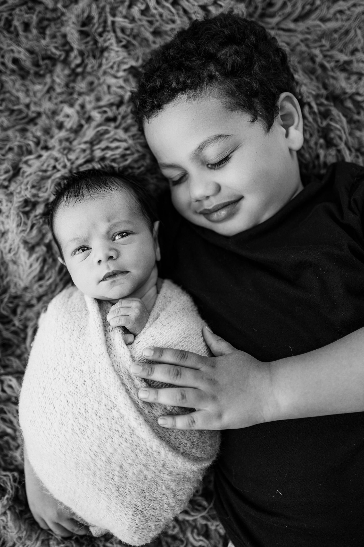 Black and white portrait of two siblings lying on a shaggy rug, with younger one wrapped in white blanket.