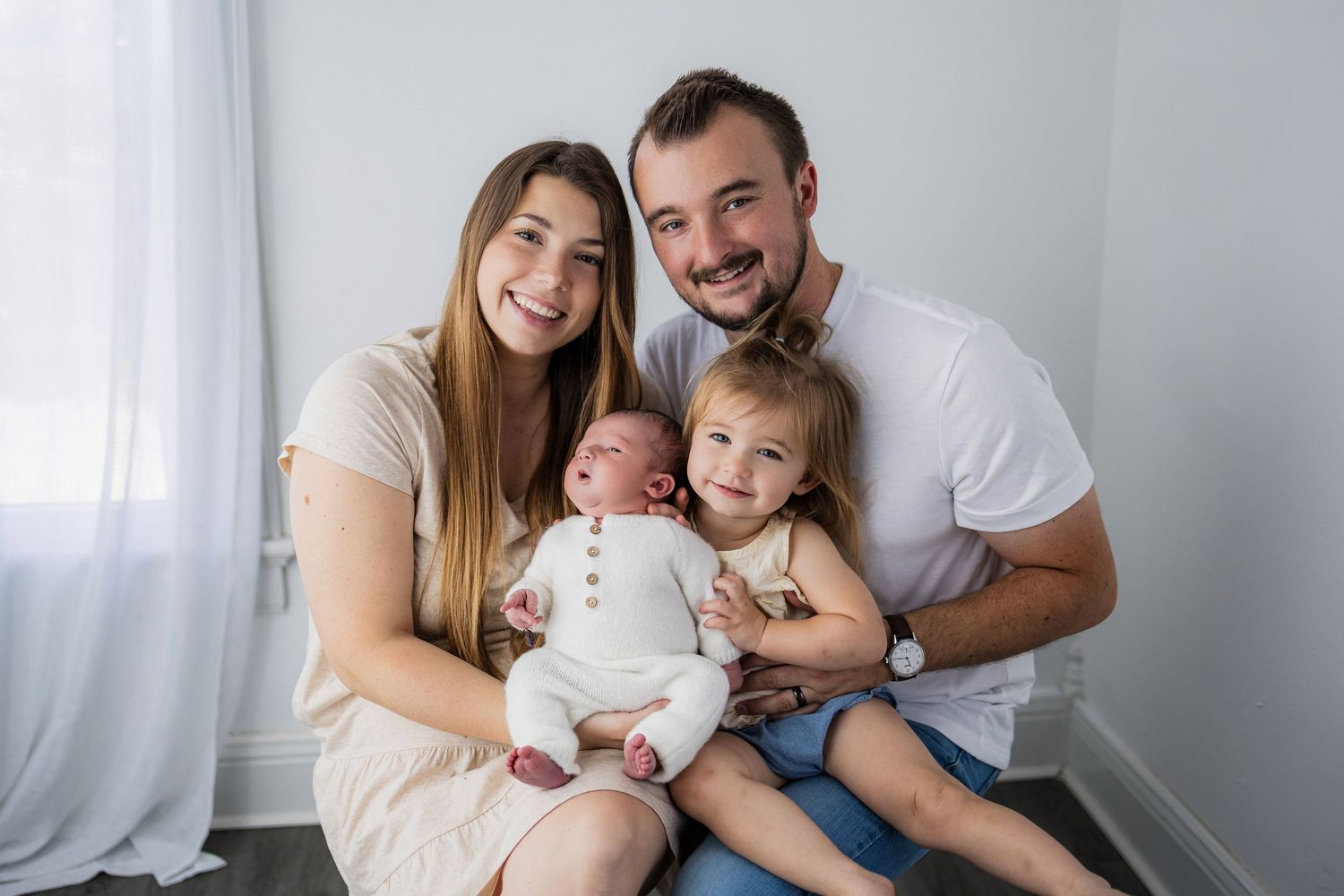 A family sits together on the floor against a white wall in their home, including a newborn baby and young child.