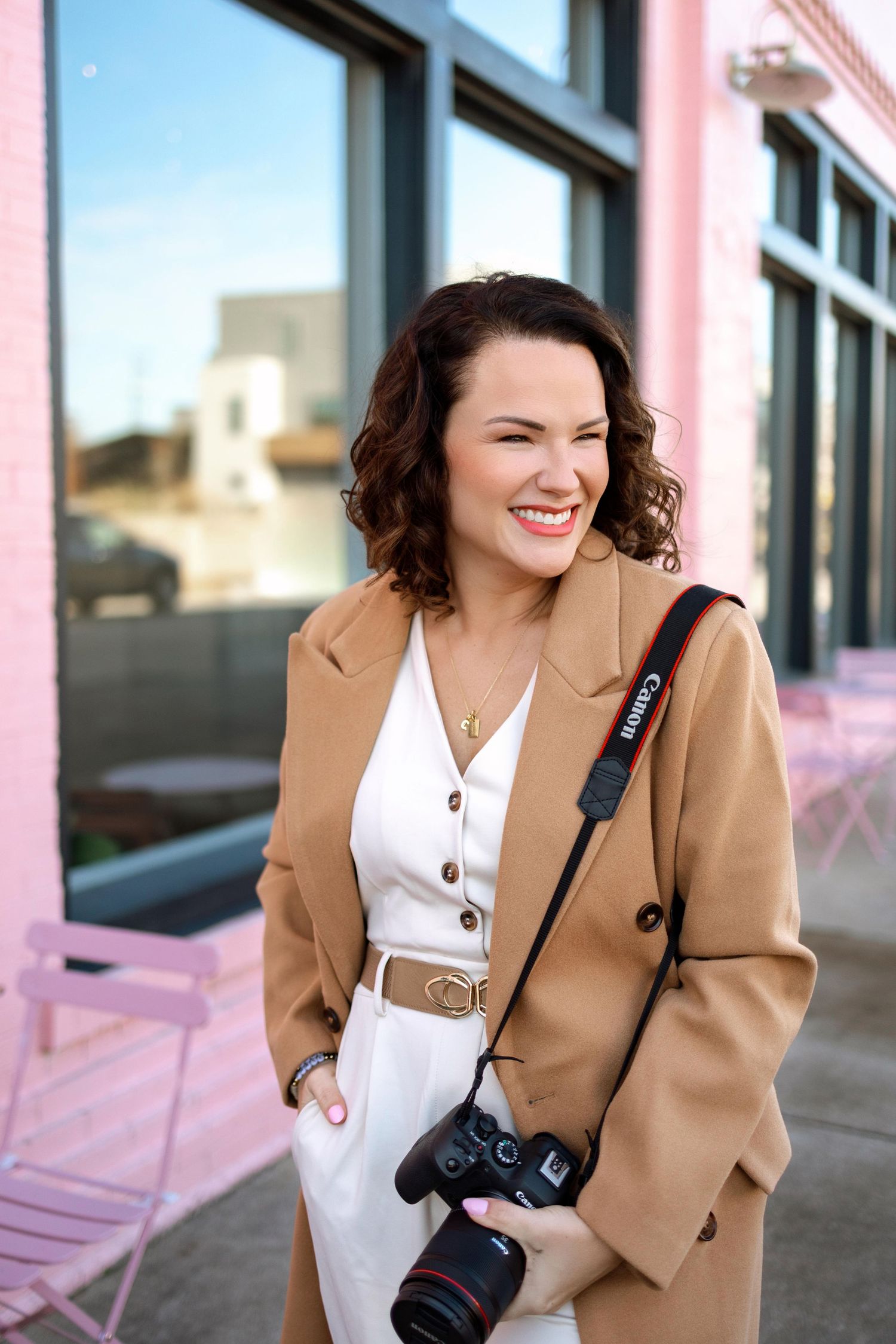 Professional photographer wearing a tan blazer and white dress with camera gear smiling outdoors by storefront windows.