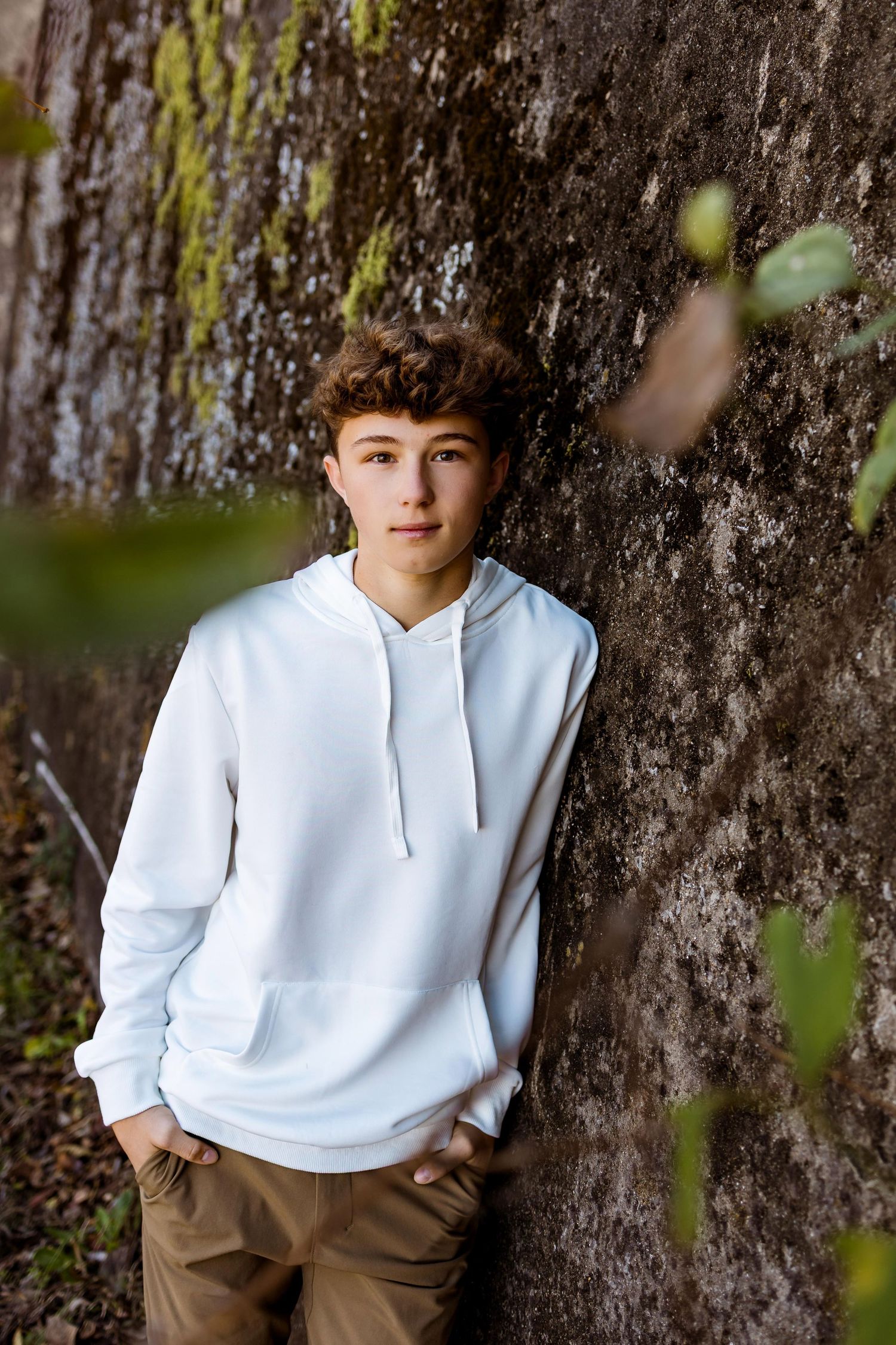 A casual portrait against a textured tree trunk background wearing a white hoodie and khaki pants.