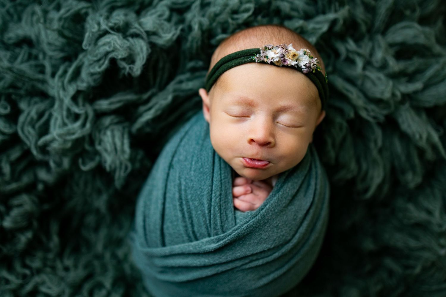 Sleeping newborn swaddled in teal fabric against a dark green textured background.