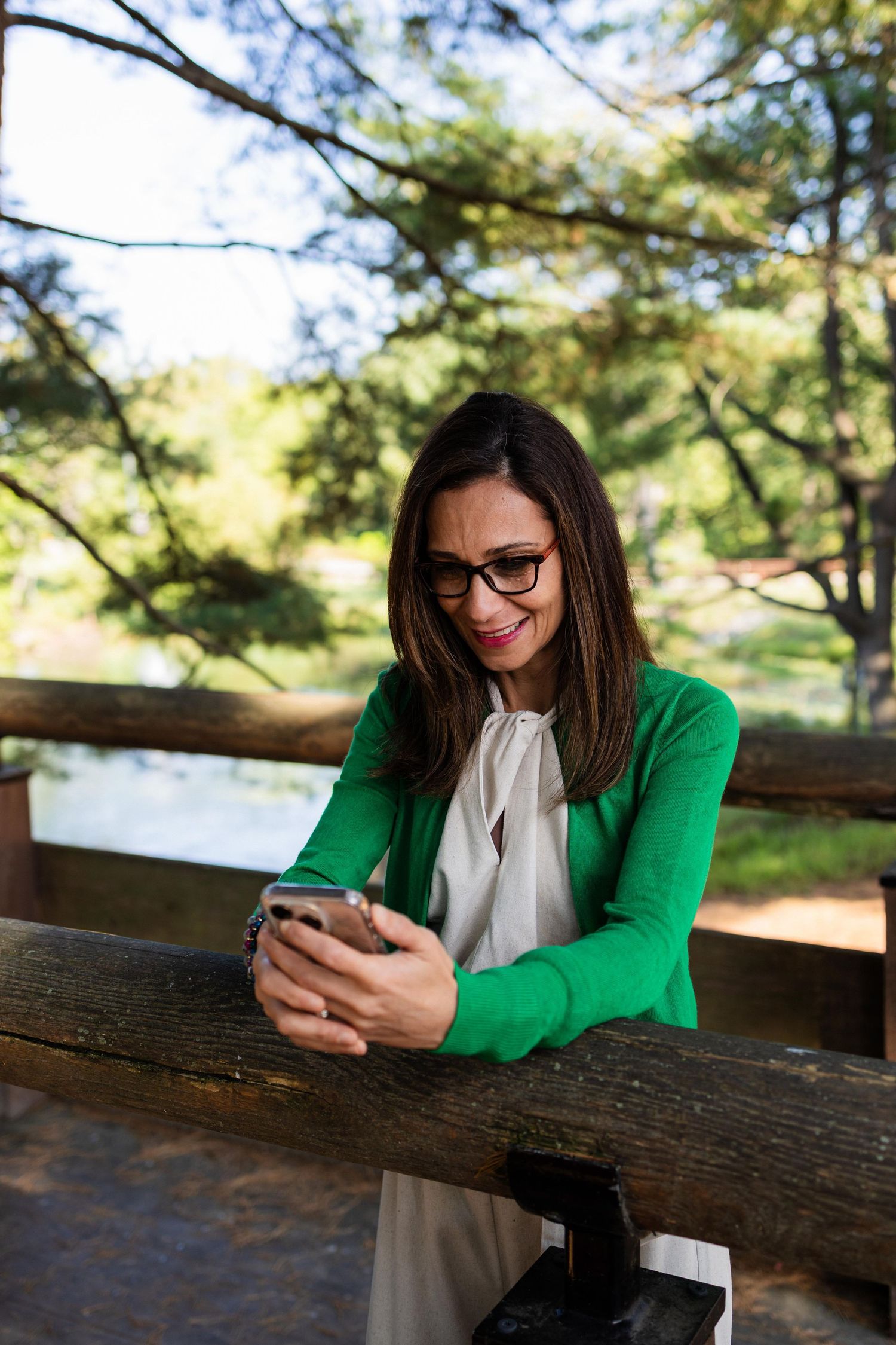 Person in green cardigan and glasses smiles while looking at phone on outdoor wooden deck.