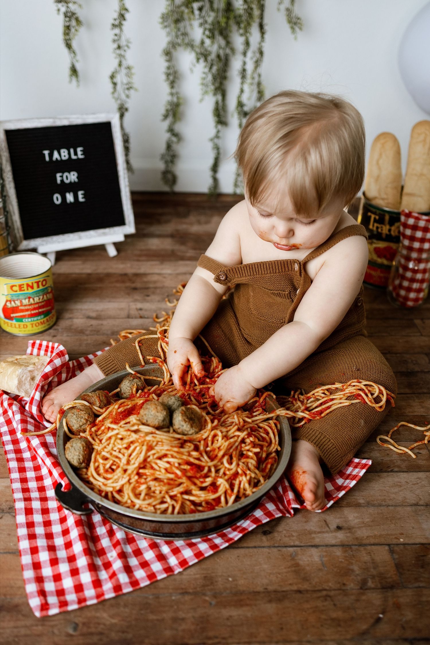 Small child in brown overalls explores plate of spaghetti and meatballs on red checkered tablecloth.