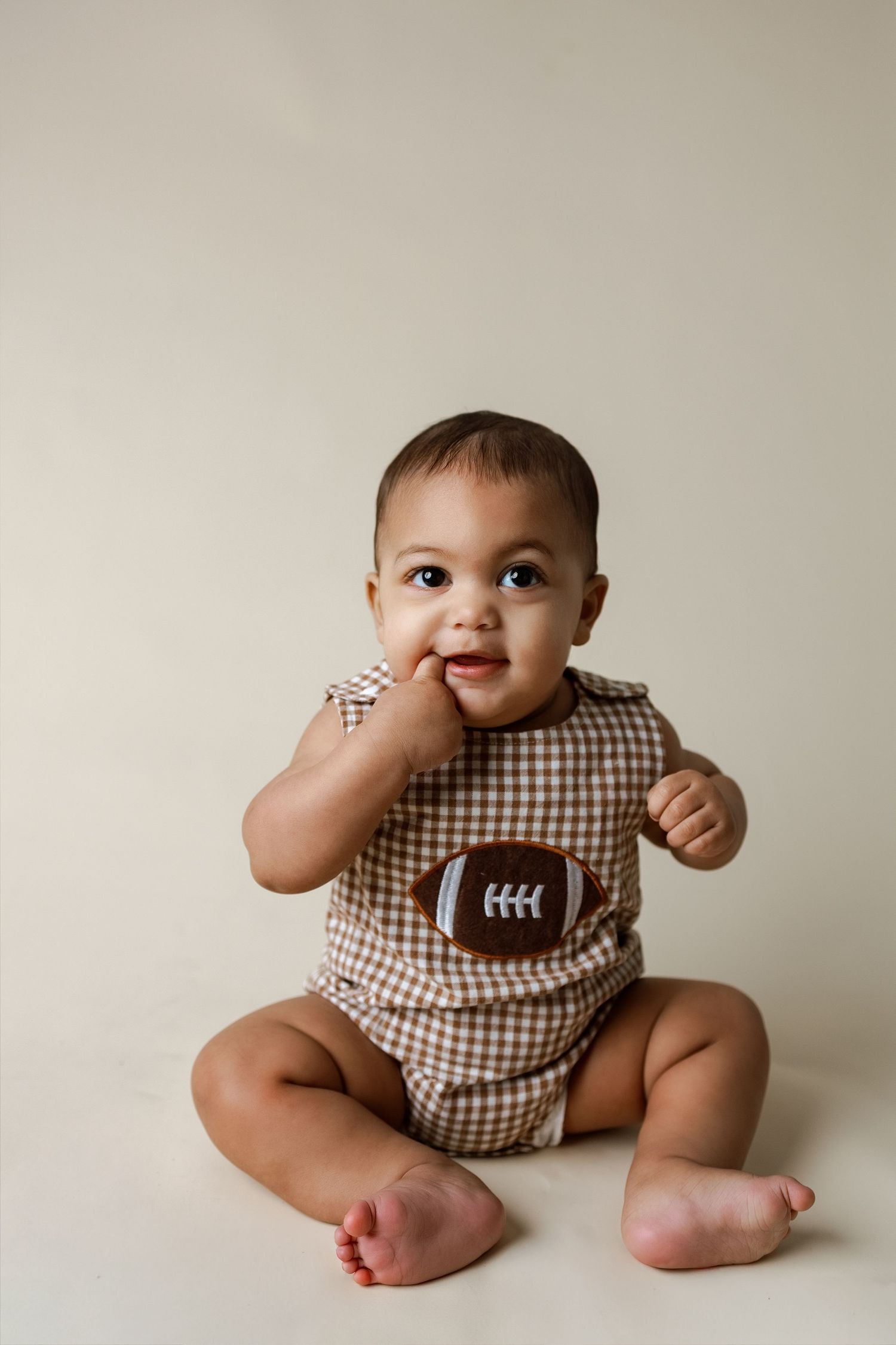 A baby wearing a brown gingham romper sits on a neutral backdrop in a series of playful poses.
