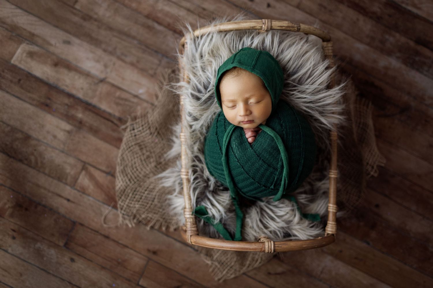 Overhead view of newborn baby wrapped in green swaddle and grey blanket on wooden floor.