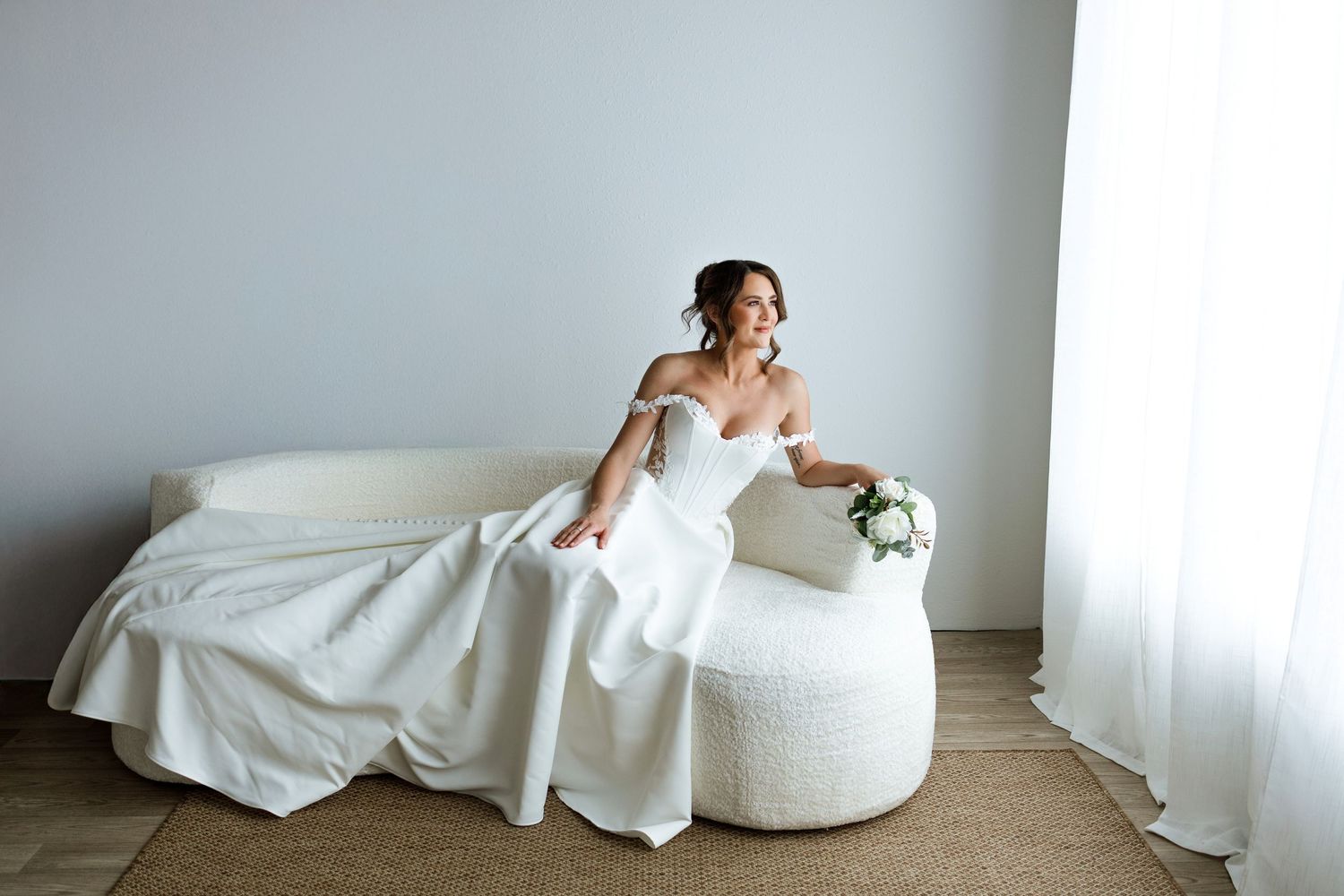 Elegant bride in white off-shoulder gown reclining on cream sofa with bouquet by window.