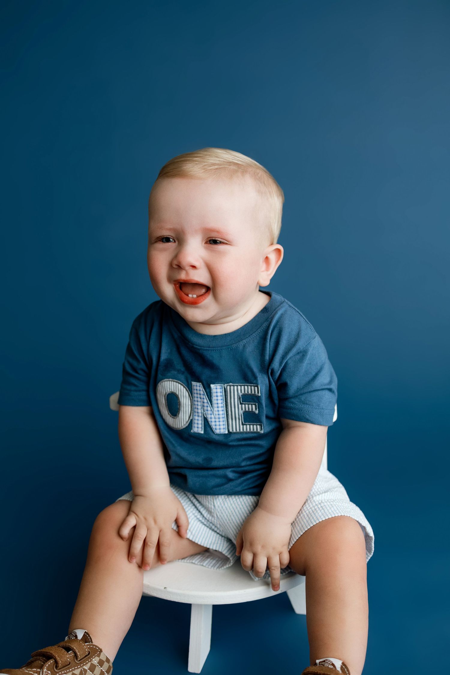 A crying toddler wearing a blue ONE shirt sits on a white stool against a blue background for his first birthday photo.