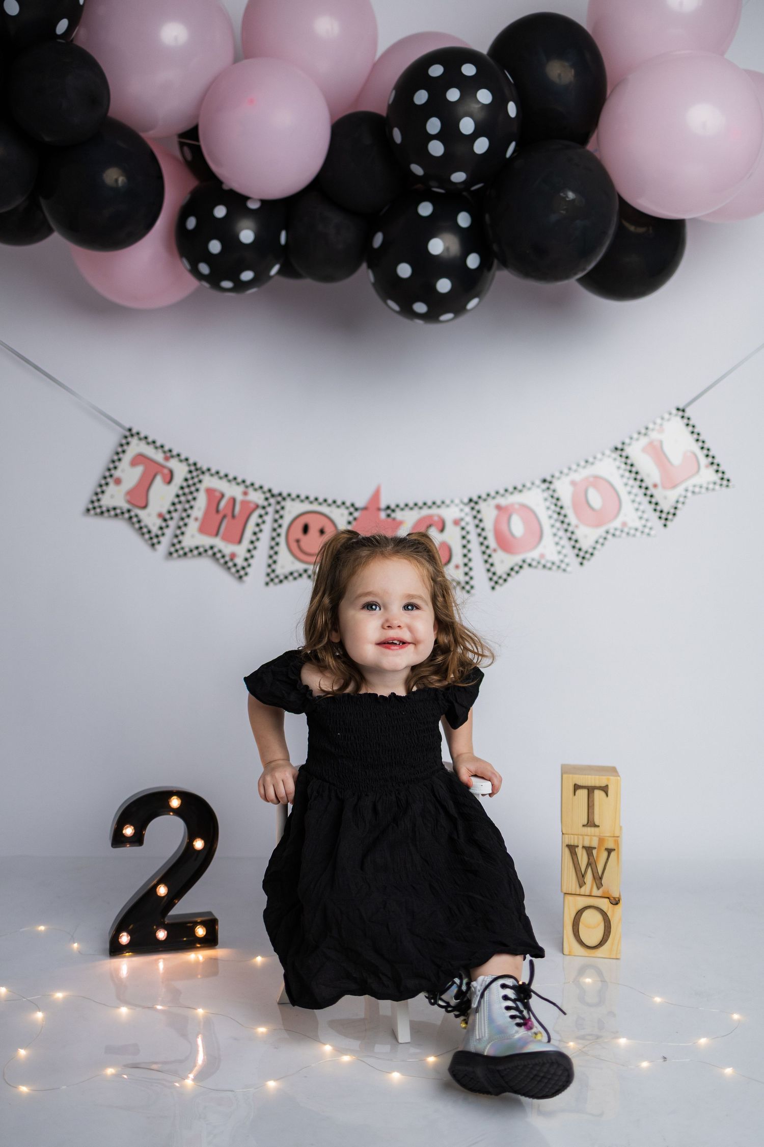 A birthday party setup with pink and black polka dot balloons, twinkling lights, and fun decorated banners in black and blush.
