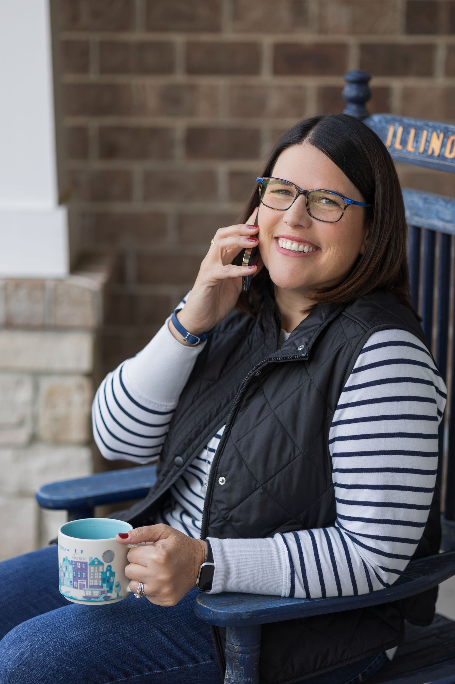 Person in a black vest and striped shirt relaxes on a porch with a blue coffee mug in hand.