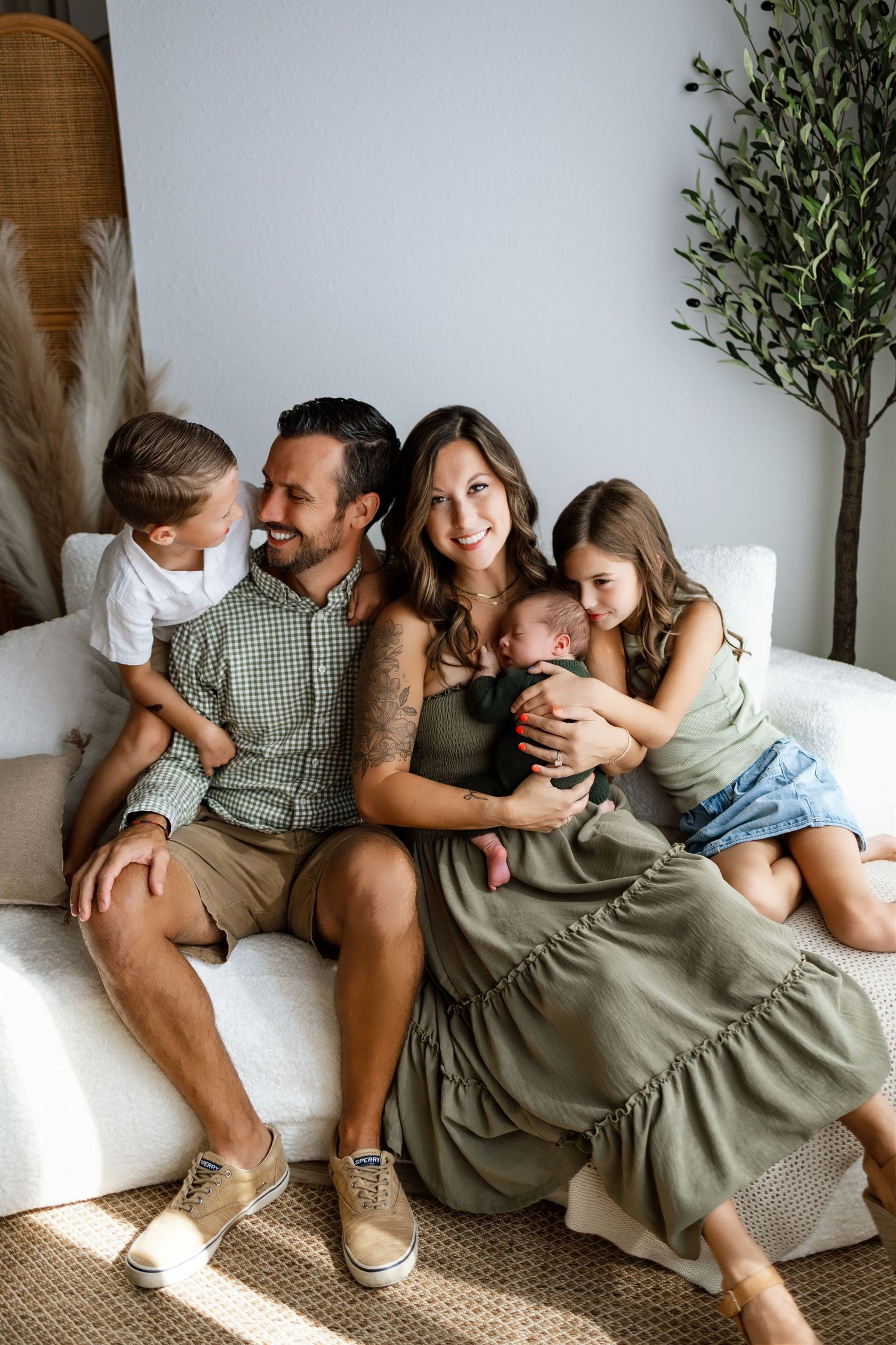 Happy family of five with parents, two young children, and a newborn baby sitting together on a white couch indoors.