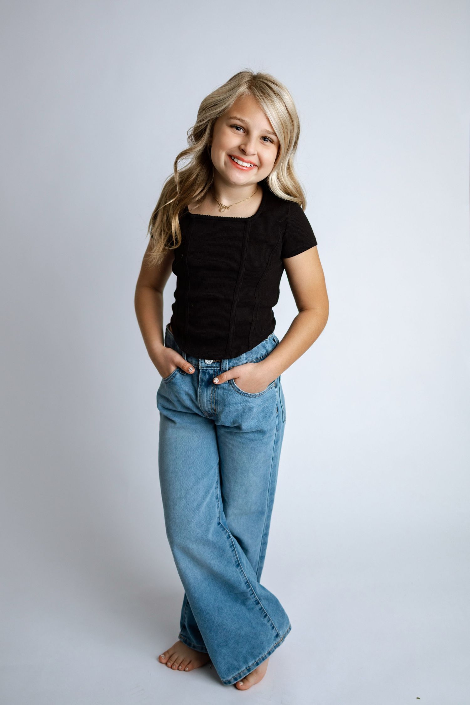 Child wearing black t-shirt and baggy blue jeans poses playfully against white background in studio setting.