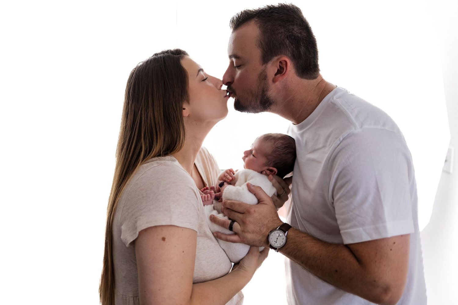 Parents in white shirts share an intimate moment while holding their newborn baby between them.