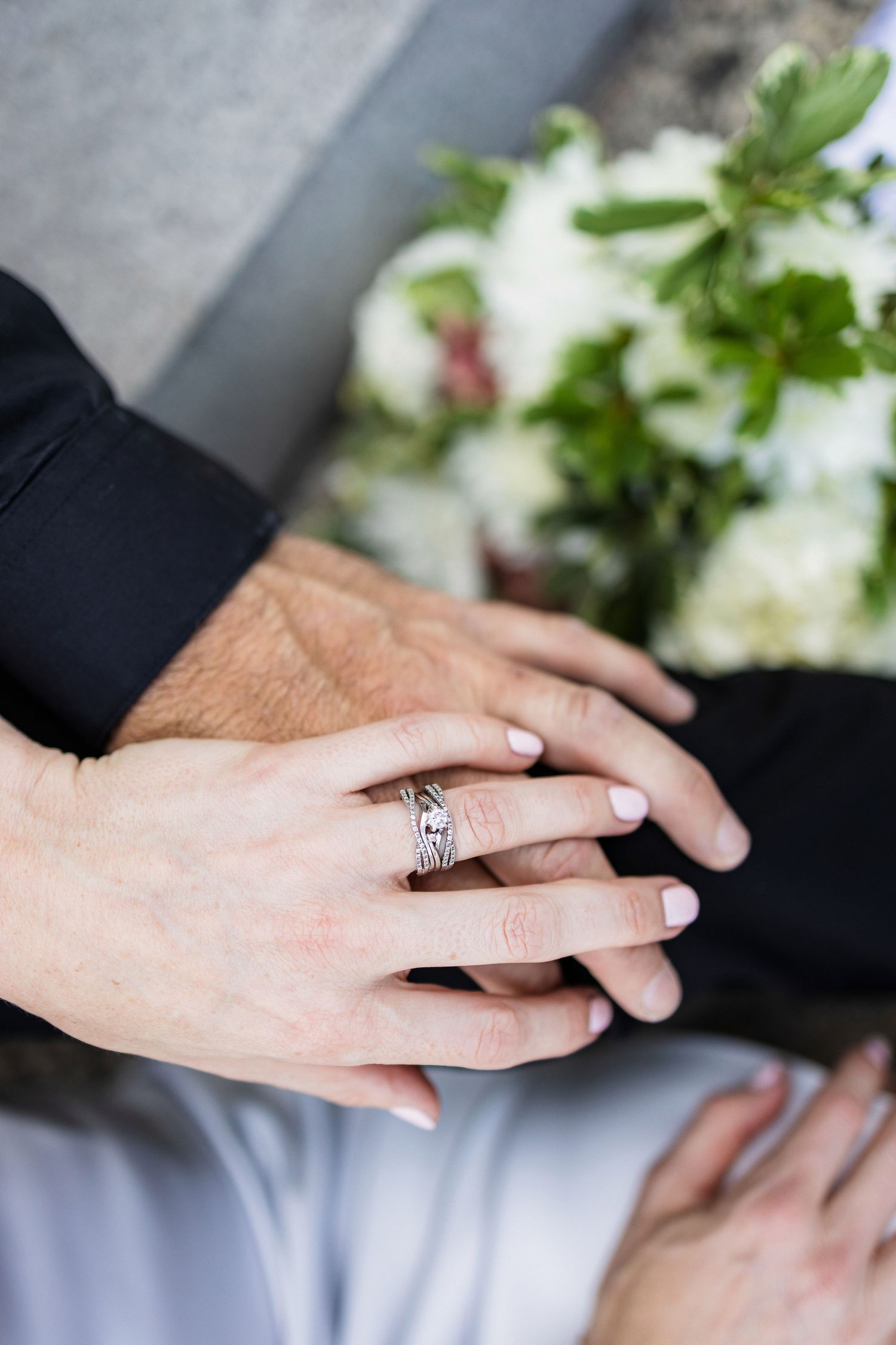 Close-up of hands showing off a detailed engagement ring with white flowers in the background.