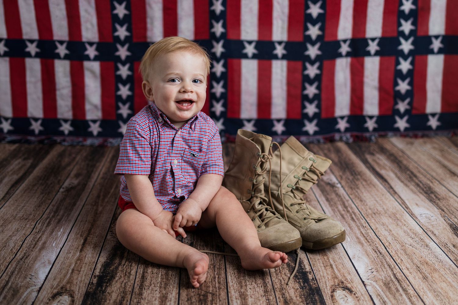 Military themed photo of a baby sitting on wooden floor next to combat boots with American flag backdrop.