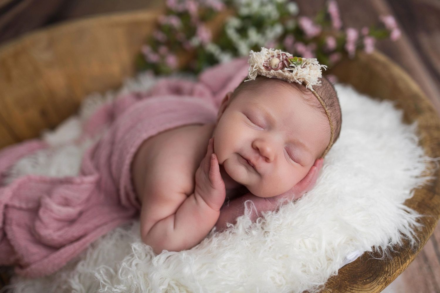 Sleeping newborn baby wearing pink wrap and tiara rests on white fur in wooden bowl.