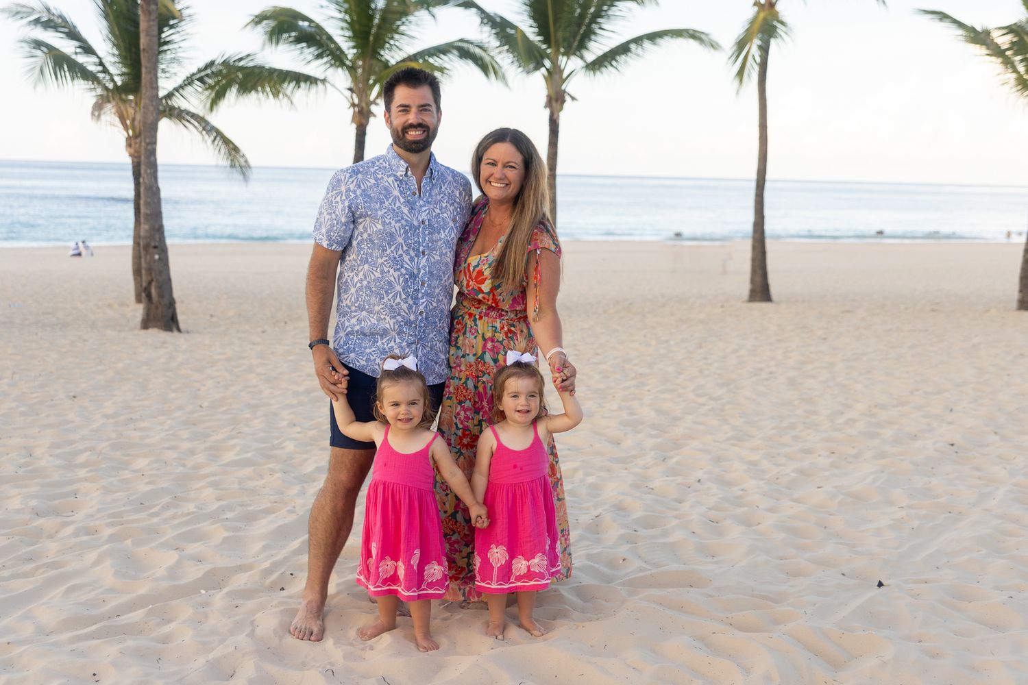 A family poses together on a tropical beach with palm trees and ocean waves in the background.