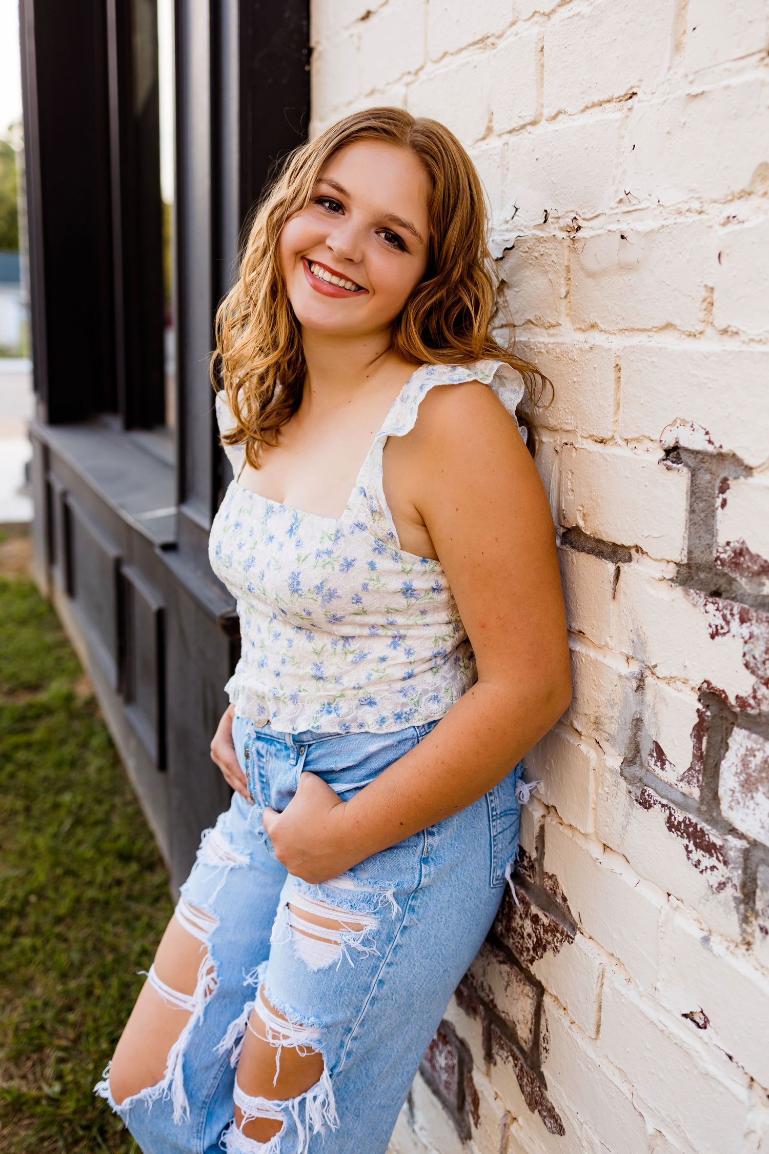 A person wearing ripped denim overalls and a floral top poses against a white brick wall in casual summer style.