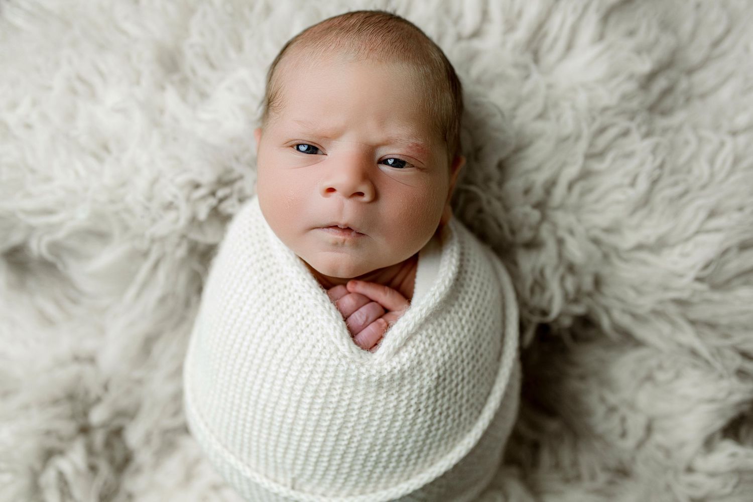 Newborn baby wrapped in soft white knit blanket lying on fluffy white fur background.