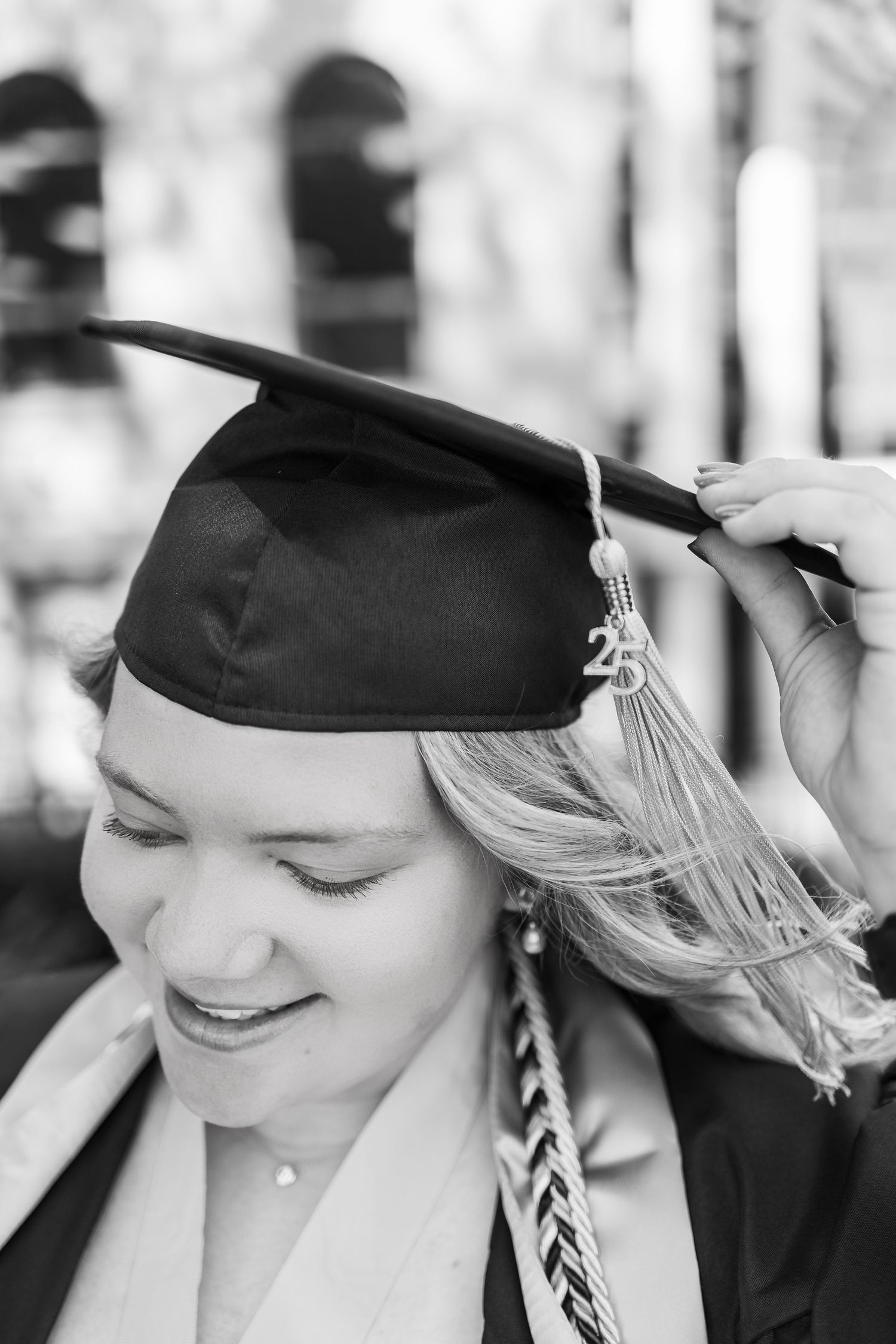 Black and white graduation portrait showing graduate in cap and gown adjusting mortarboard with silver tassel.