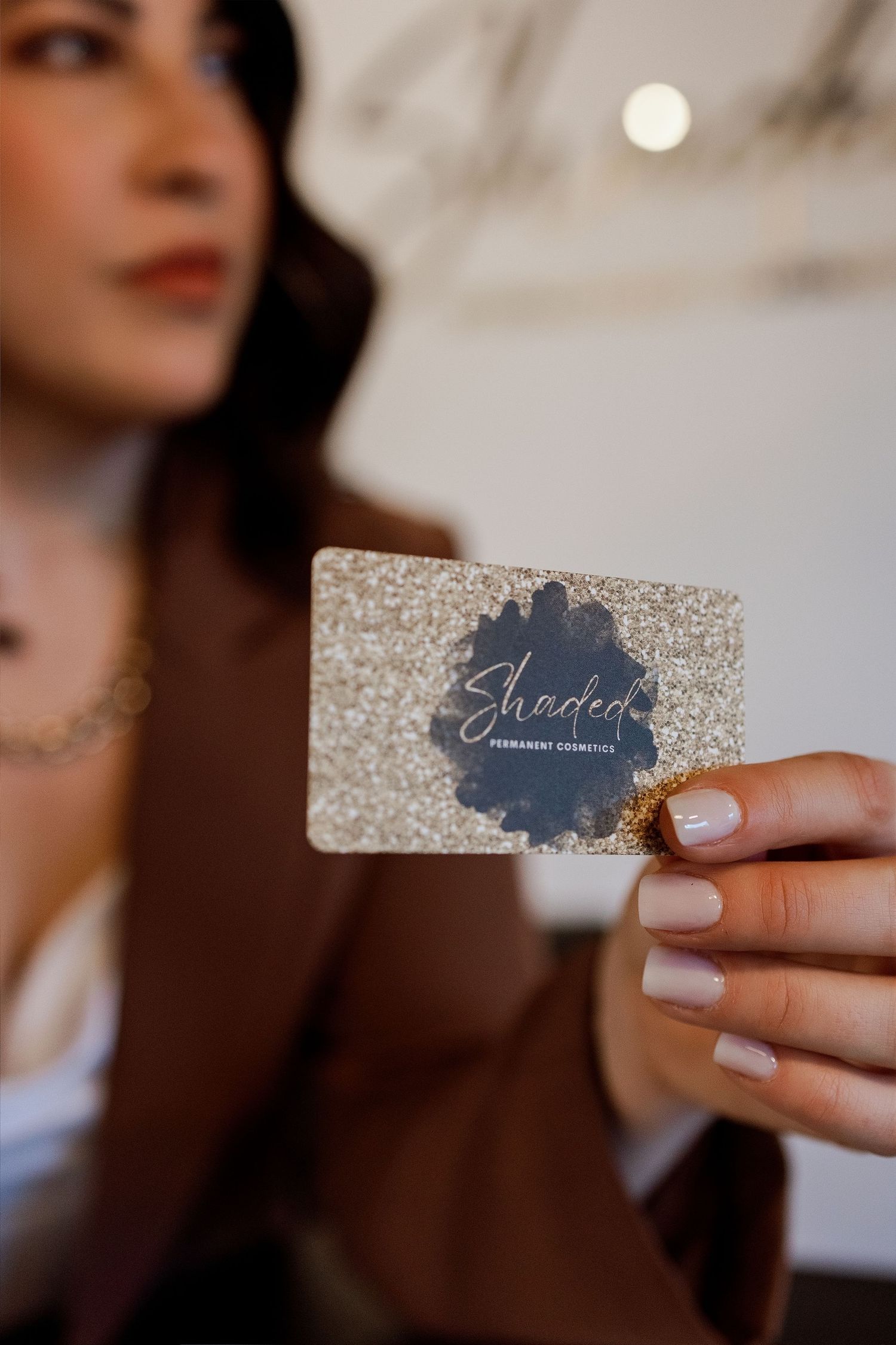 A hand holds a silver glitter business card with elegant text design against a soft blurred background.