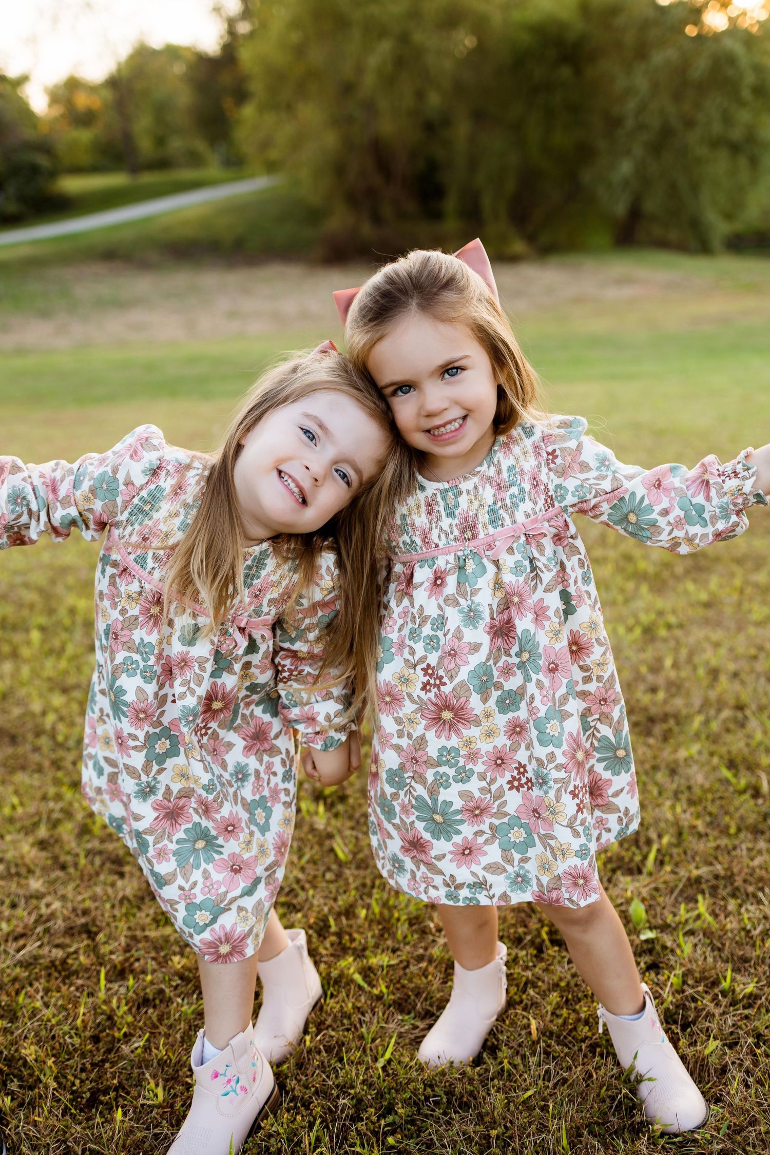 Two children in matching floral dresses pose playfully on grass during golden hour sunset.
