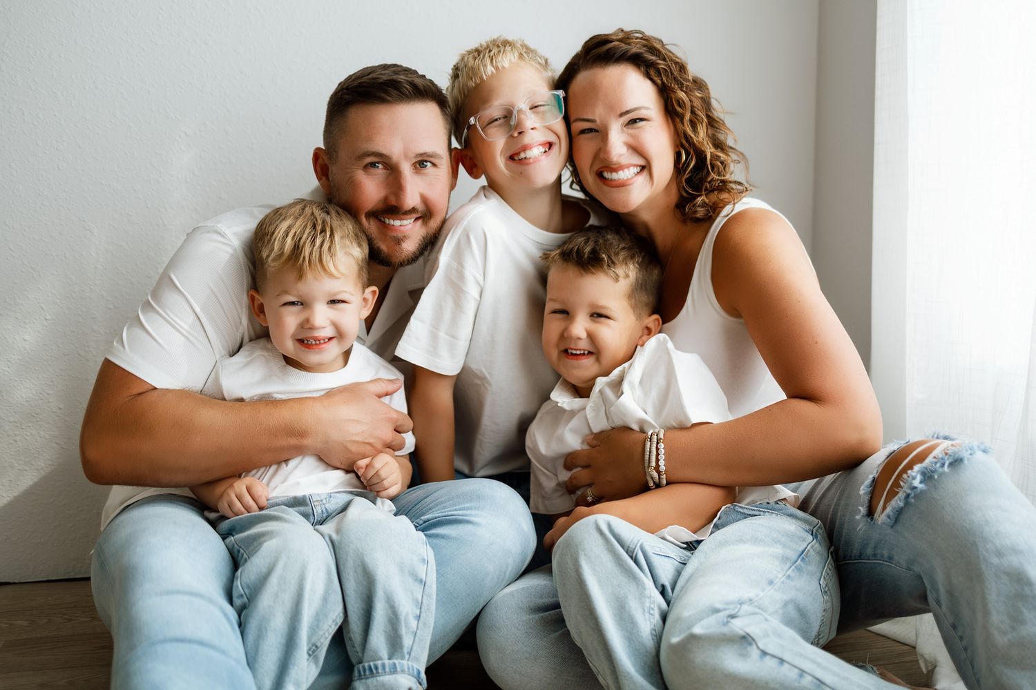 Happy family of five posing together, parents and three young boys wearing white shirts and jeans in bright studio.