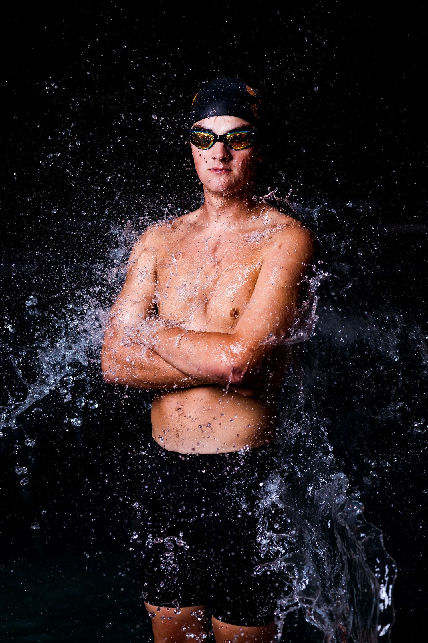 A competitive swimmer wearing goggles stands in dramatic black backdrop with water splashing around them in action sequence.