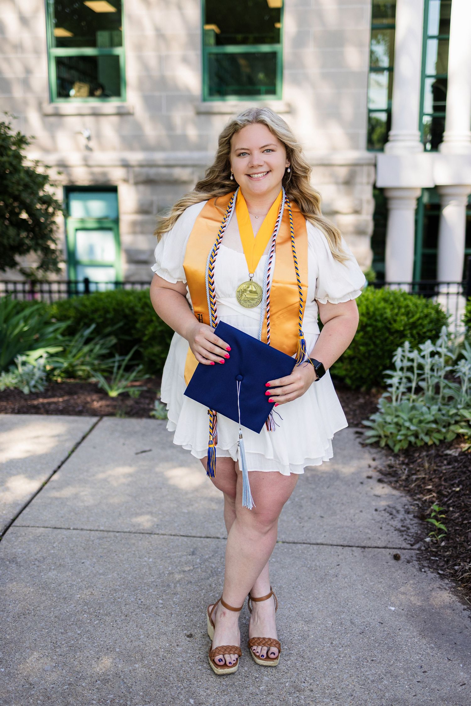 Graduate in white dress and orange stoles poses outdoors on graduation day at campus with diploma holder.