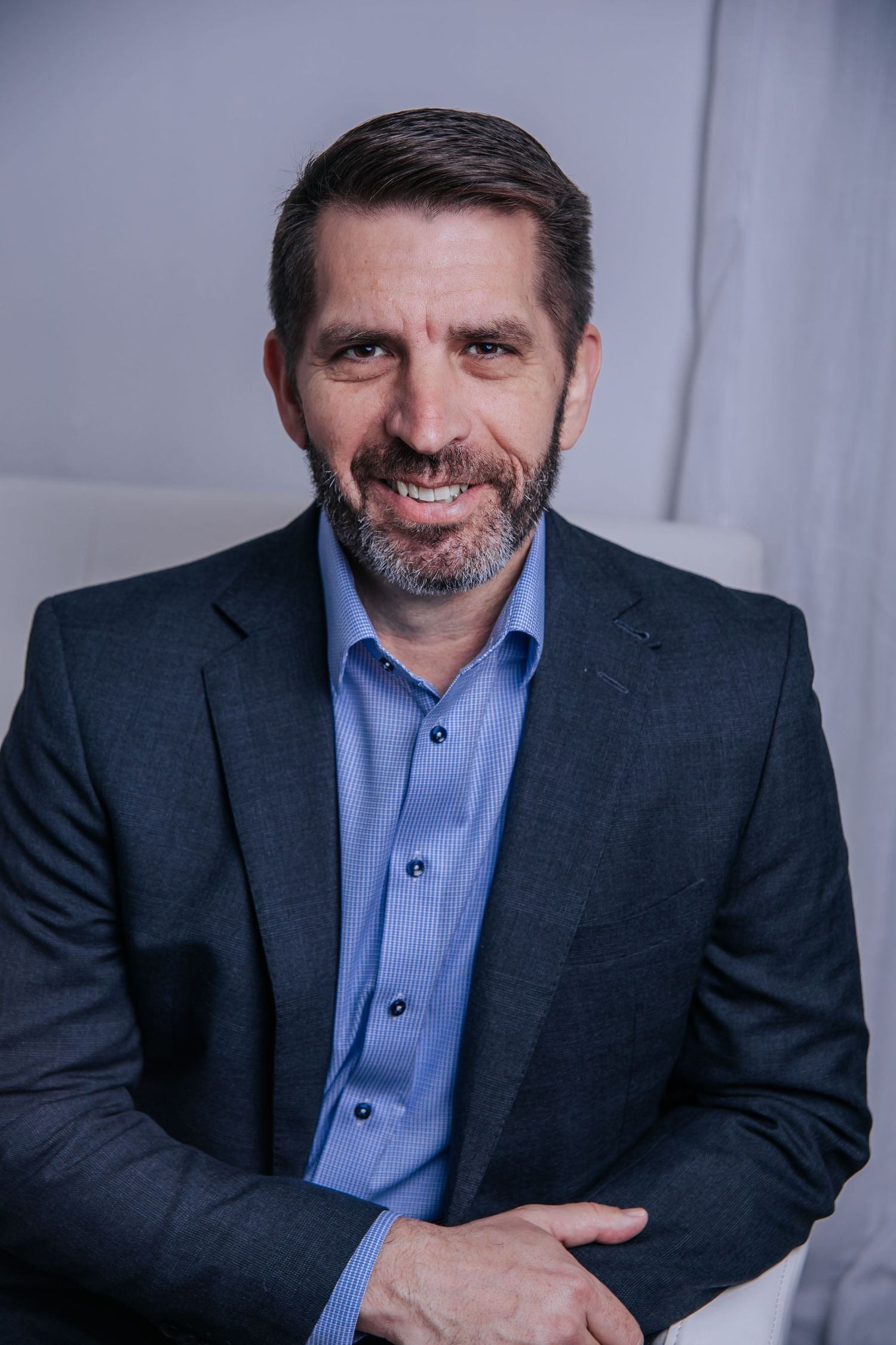 Professional headshot of person in dark suit and blue button-up shirt against grey background.
