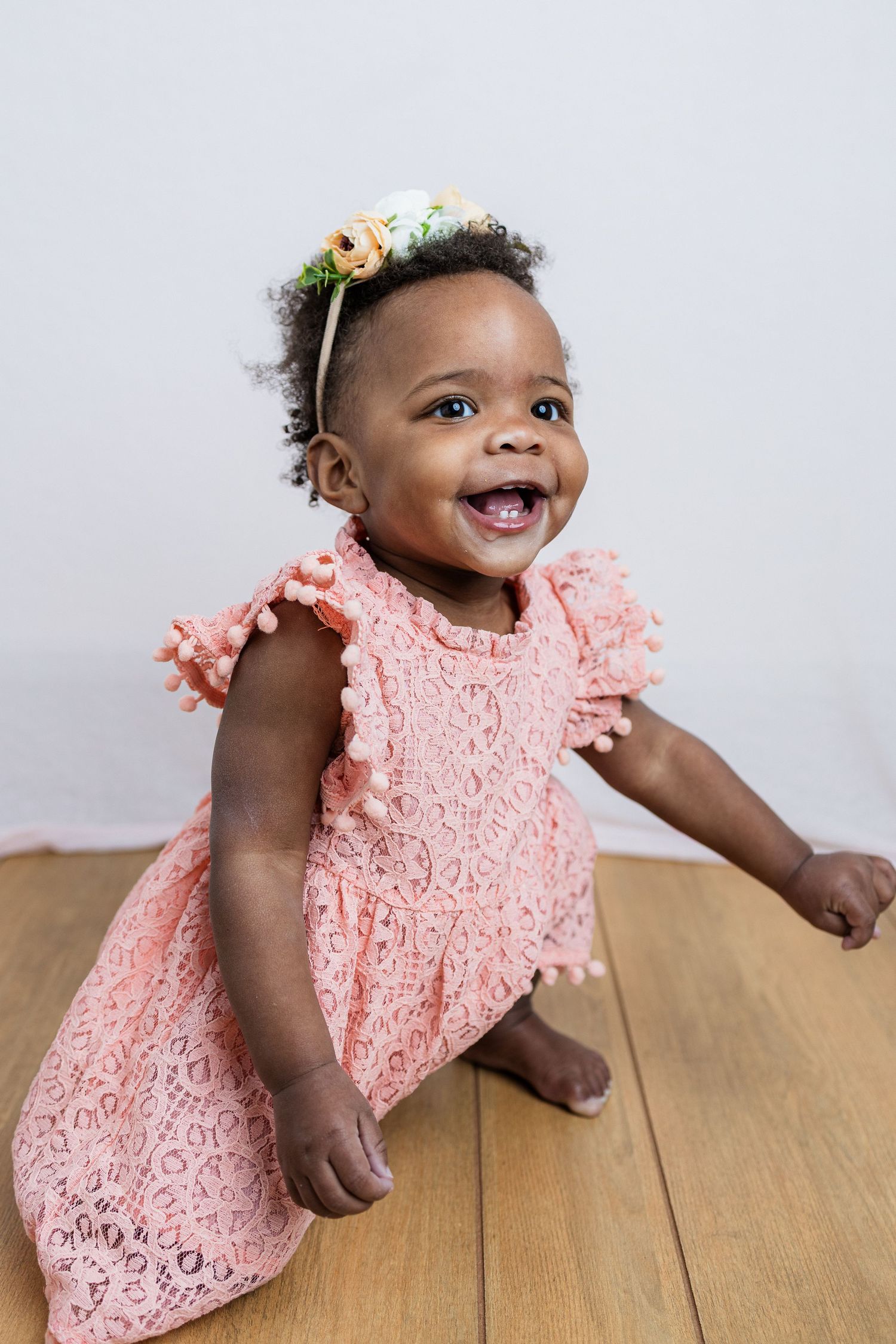 Excited toddler wearing pink lace dress and floral headband standing on hardwood floor.