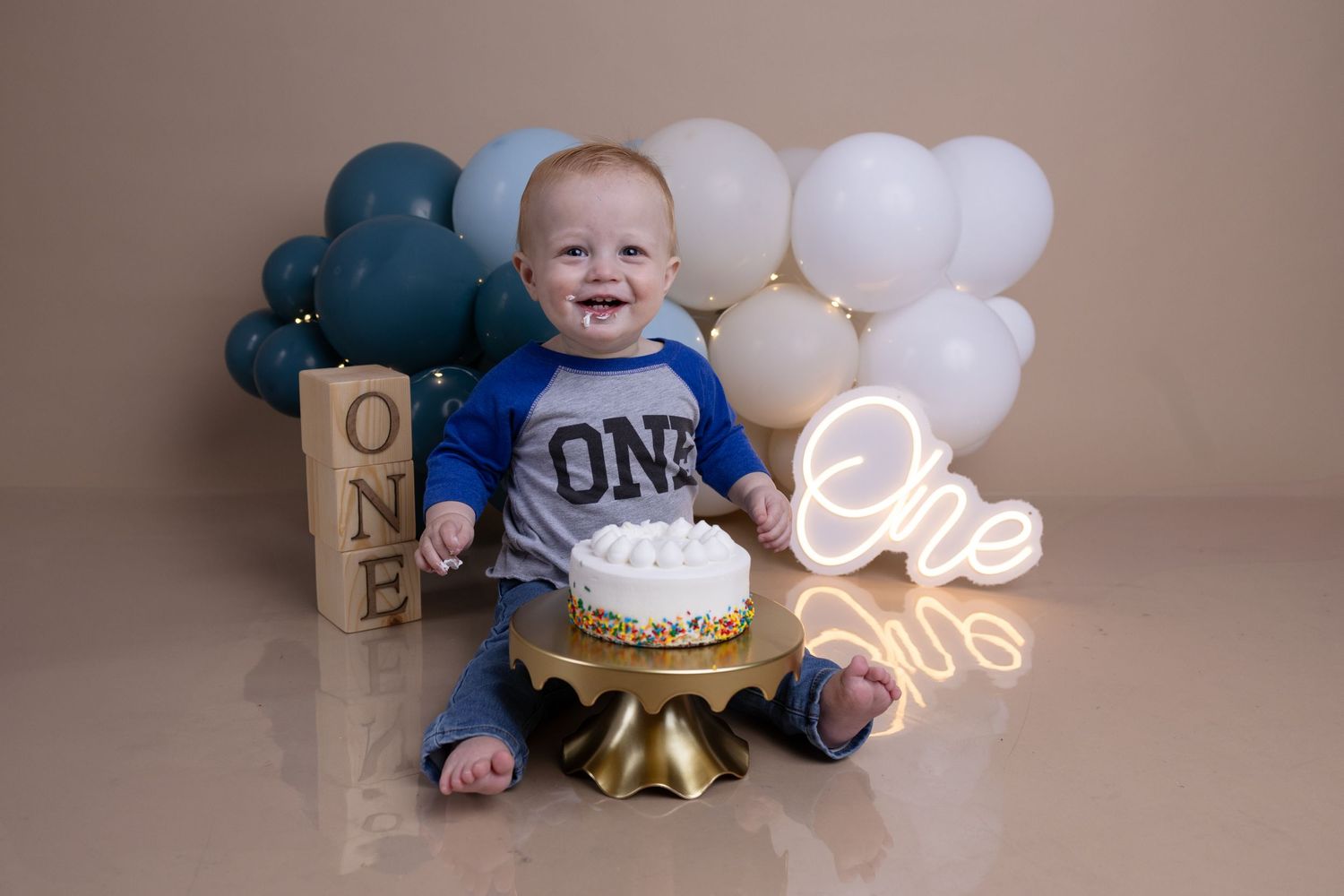 Baby in blue ONE shirt with first birthday cake setup and balloons.