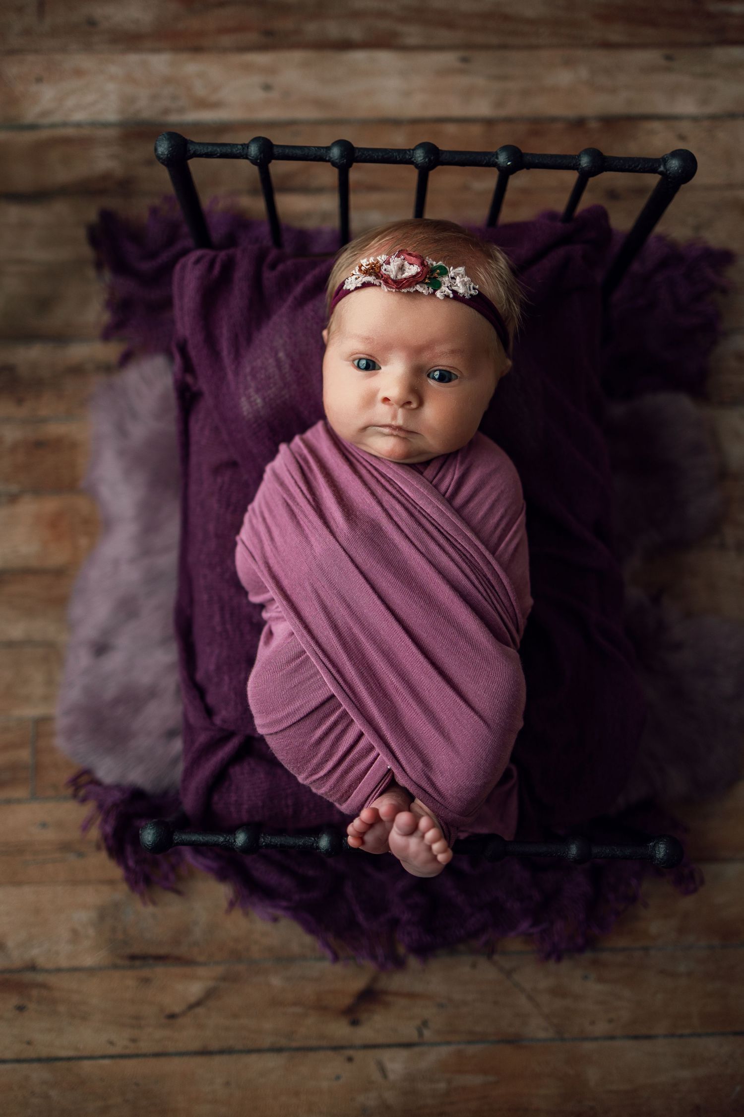 A newborn swaddled in a purple wrap lies on a dark purple blanket against a rustic wooden backdrop.