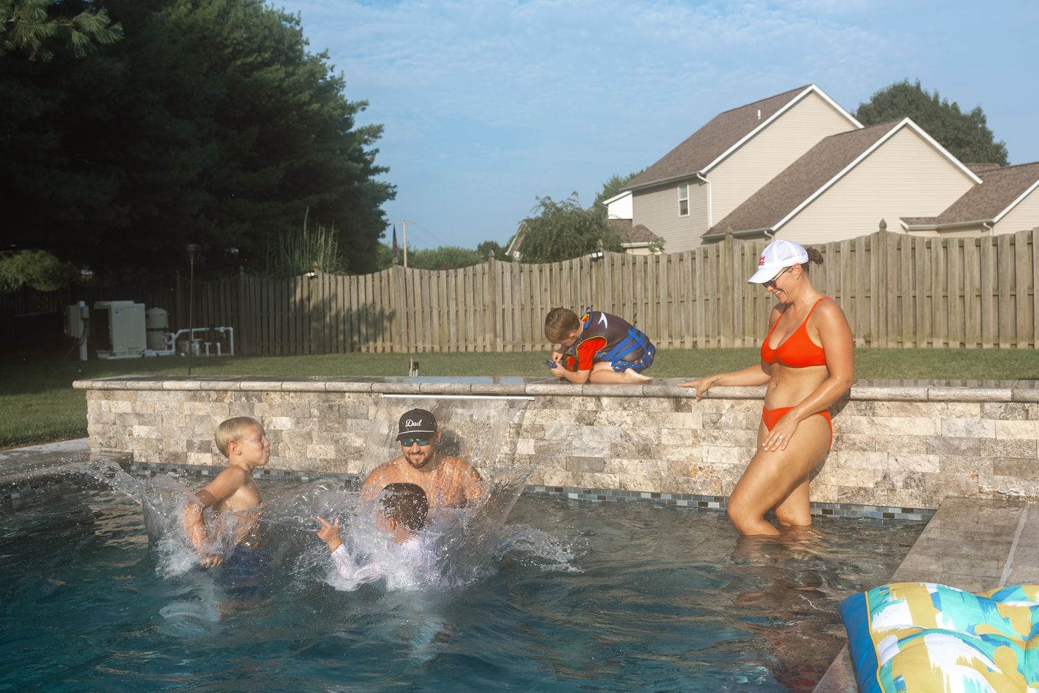 Family enjoying a backyard swimming pool on a sunny day, with children splashing and a woman in an orange bikini.