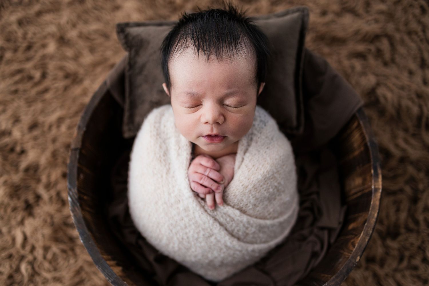 Newborn baby peacefully sleeping while swaddled in white wrap and nestled in round wooden bowl on brown backdrop.