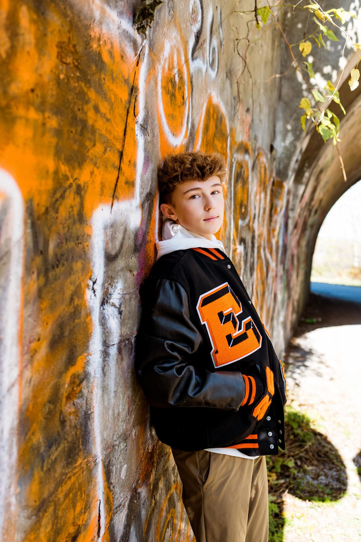 A letterman jacket in black and orange leans against a graffiti-covered concrete wall in an outdoor setting.
