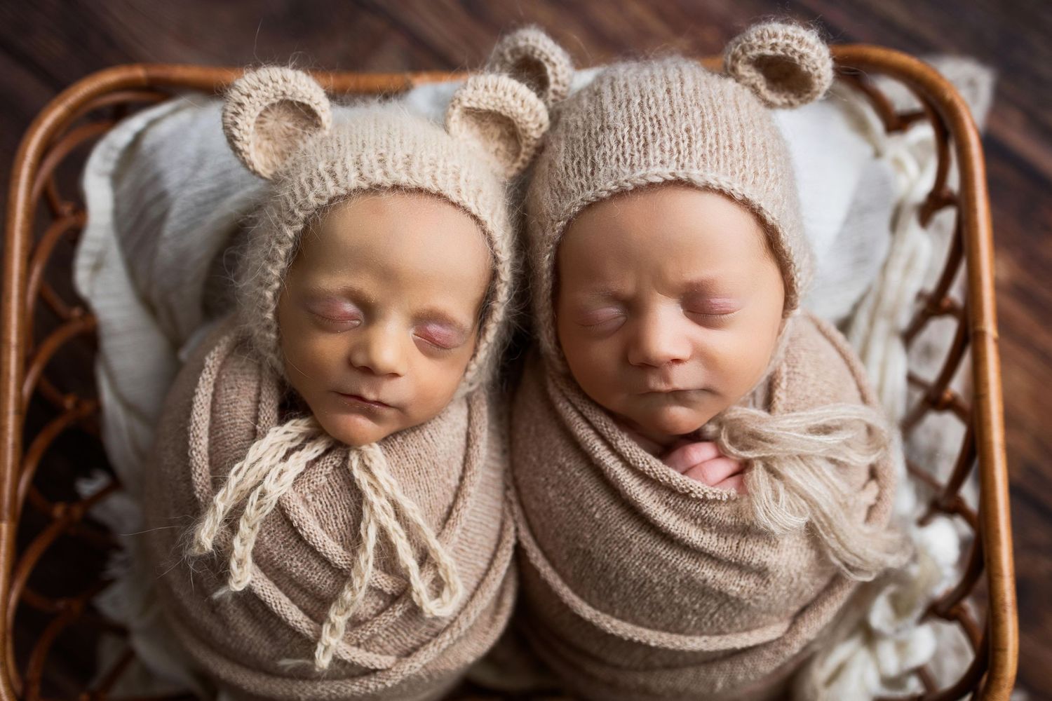 Adorable sleeping newborn twins wearing matching knitted bonnets and wraps lie together in a cozy basket.