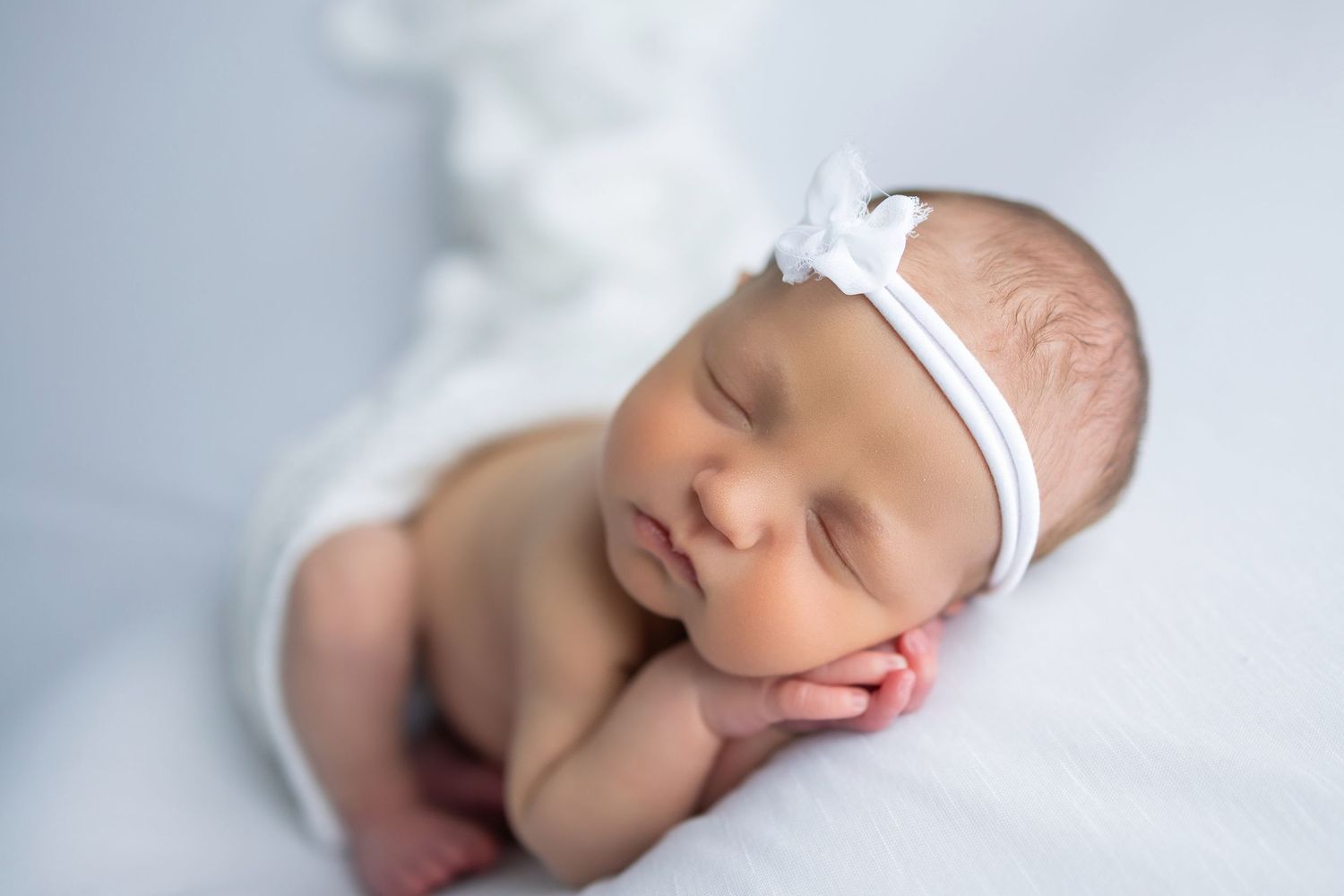 Peaceful sleeping newborn with white headband resting head on hands against light background.